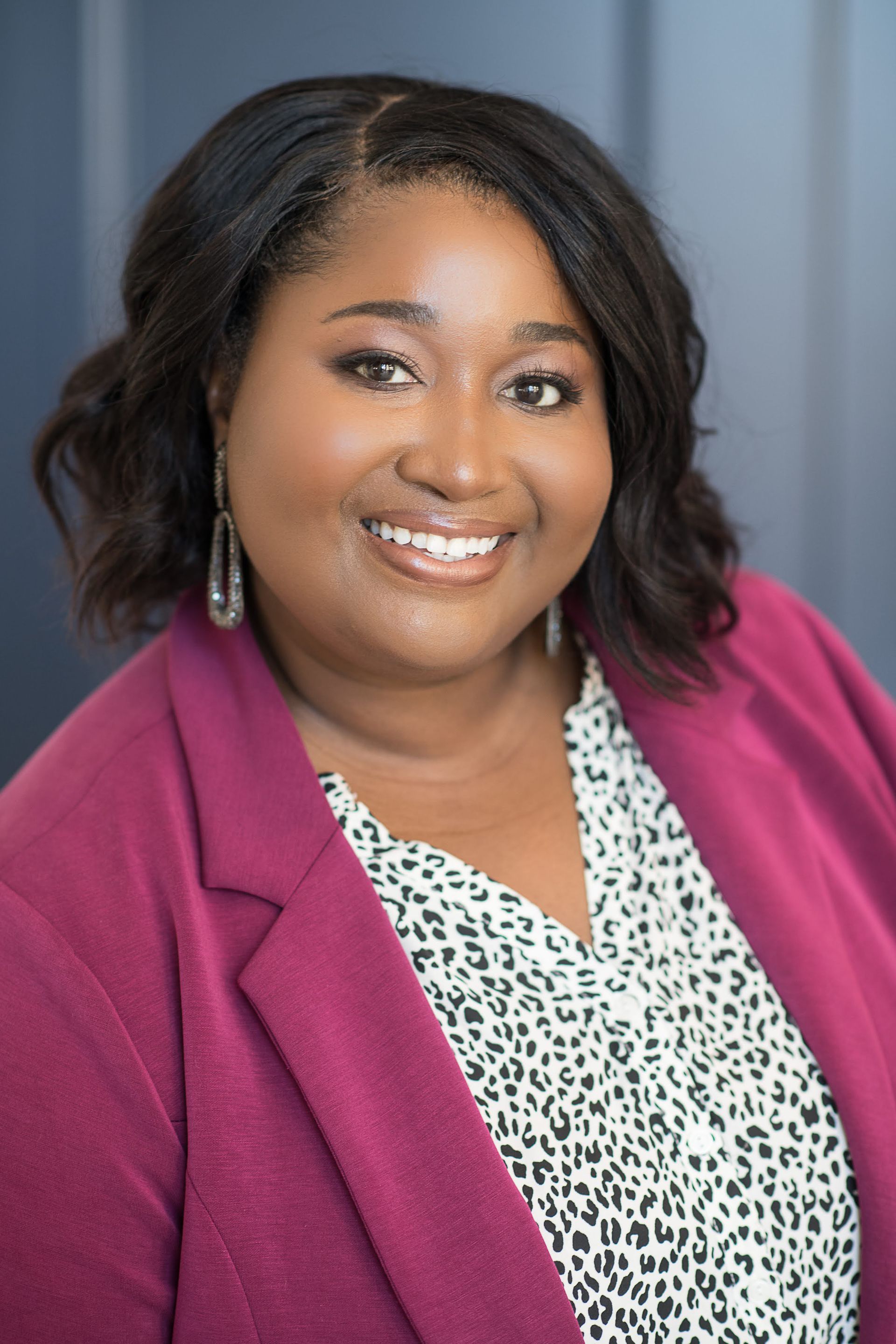 Smiling woman in a magenta blazer and patterned blouse against a gray studio backdrop