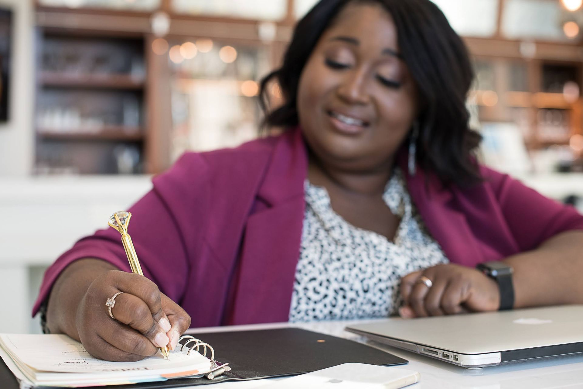 Woman in a magenta blazer writing in a notebook beside a laptop in a bright office