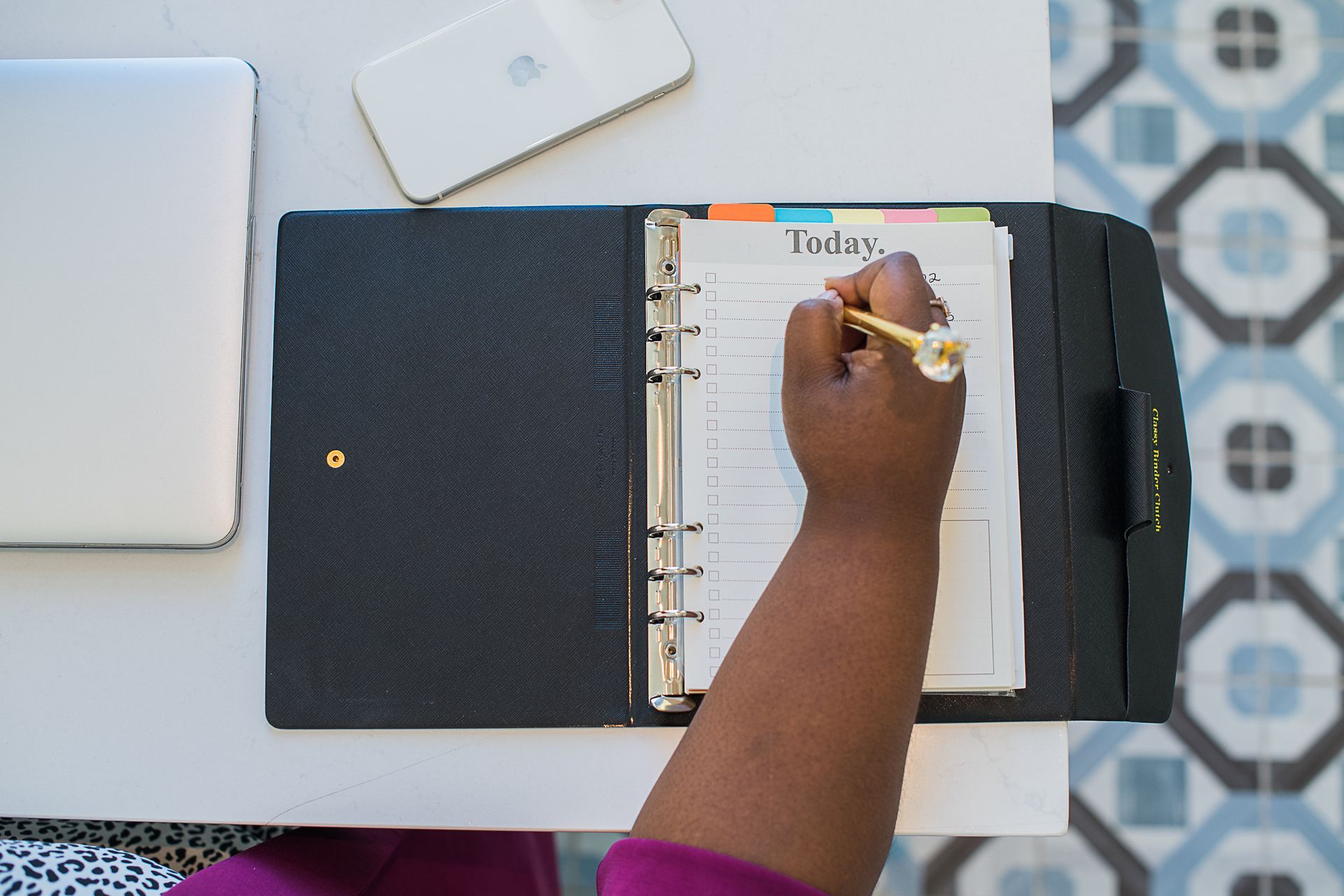 Hand writing in a black binder notebook on a white desk with a laptop and tablet nearby