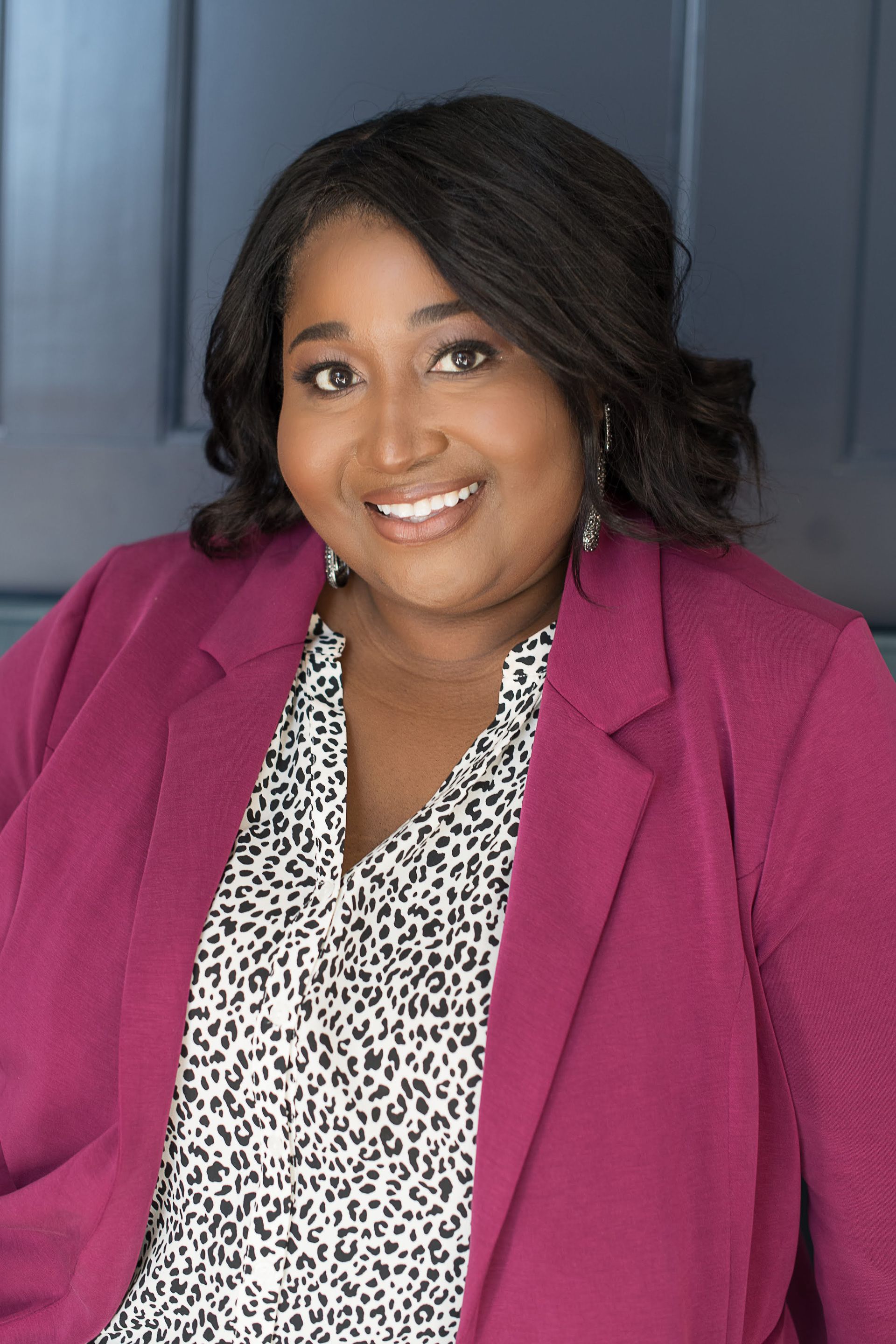Smiling person in a magenta blazer and patterned blouse against a dark studio background