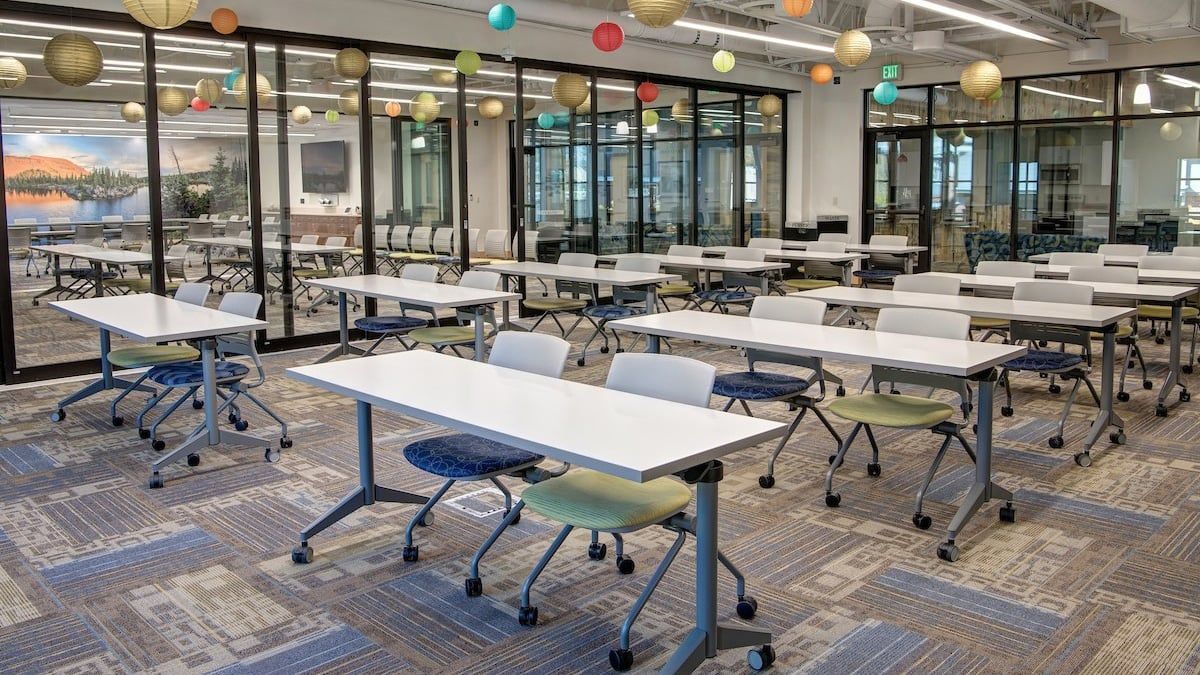 Classroom with rectangular tables and rolling chairs, glass walls, and colorful decor.
