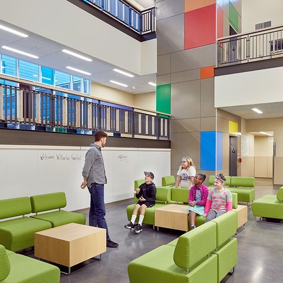Teacher talks to students seated on green couches in a brightly colored, modern school lobby.