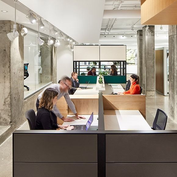 Office interior with people at desks, working, with concrete columns and neutral tones.
