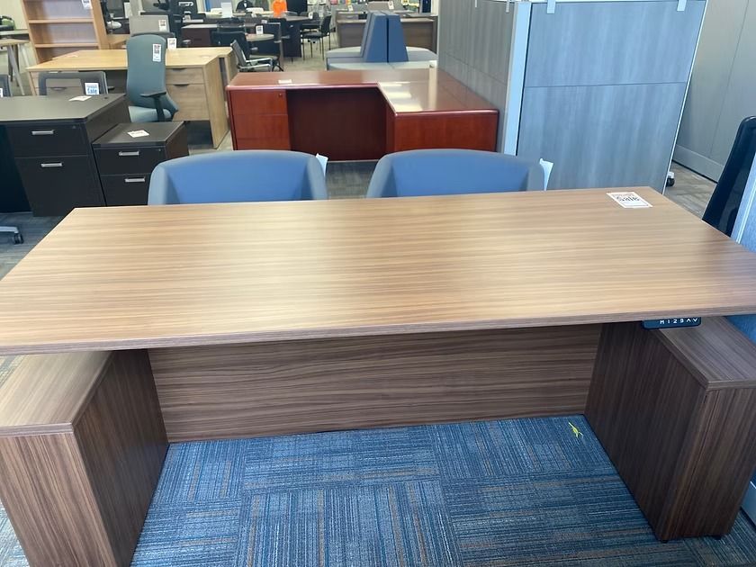 Wooden office desk with two gray chairs; other office furniture in background.