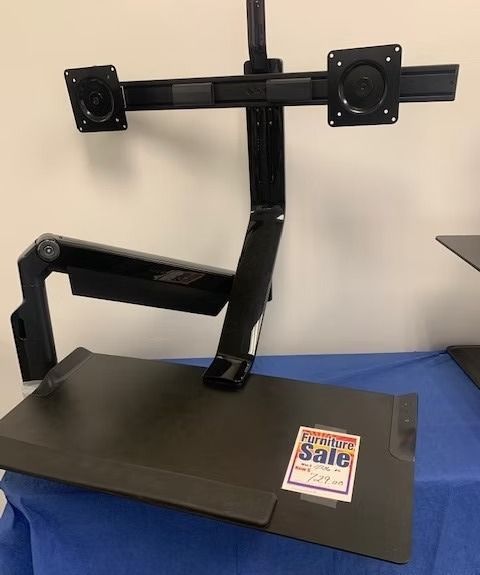 Black adjustable desk with dual monitor mount, on a blue-covered table, for sale.