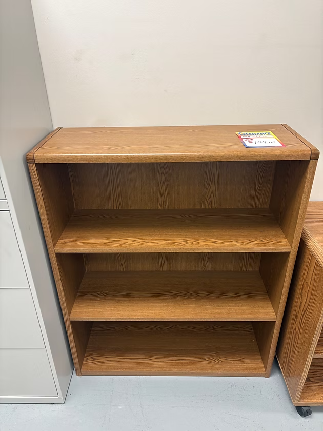 A wooden bookcase with three shelves in a room with a light-colored wall.