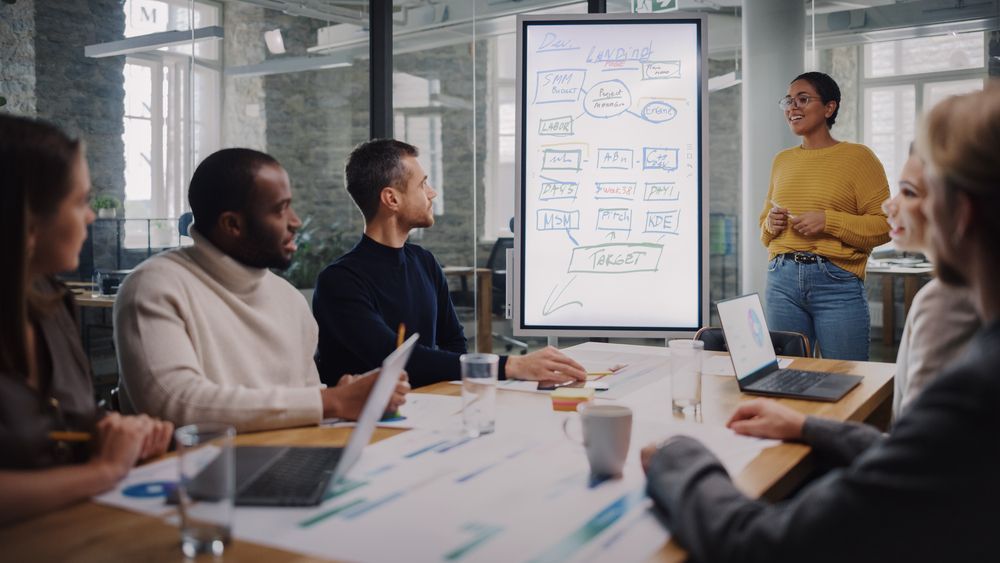 Group of people in a meeting. A woman presents a diagram on a screen. Modern office setting.