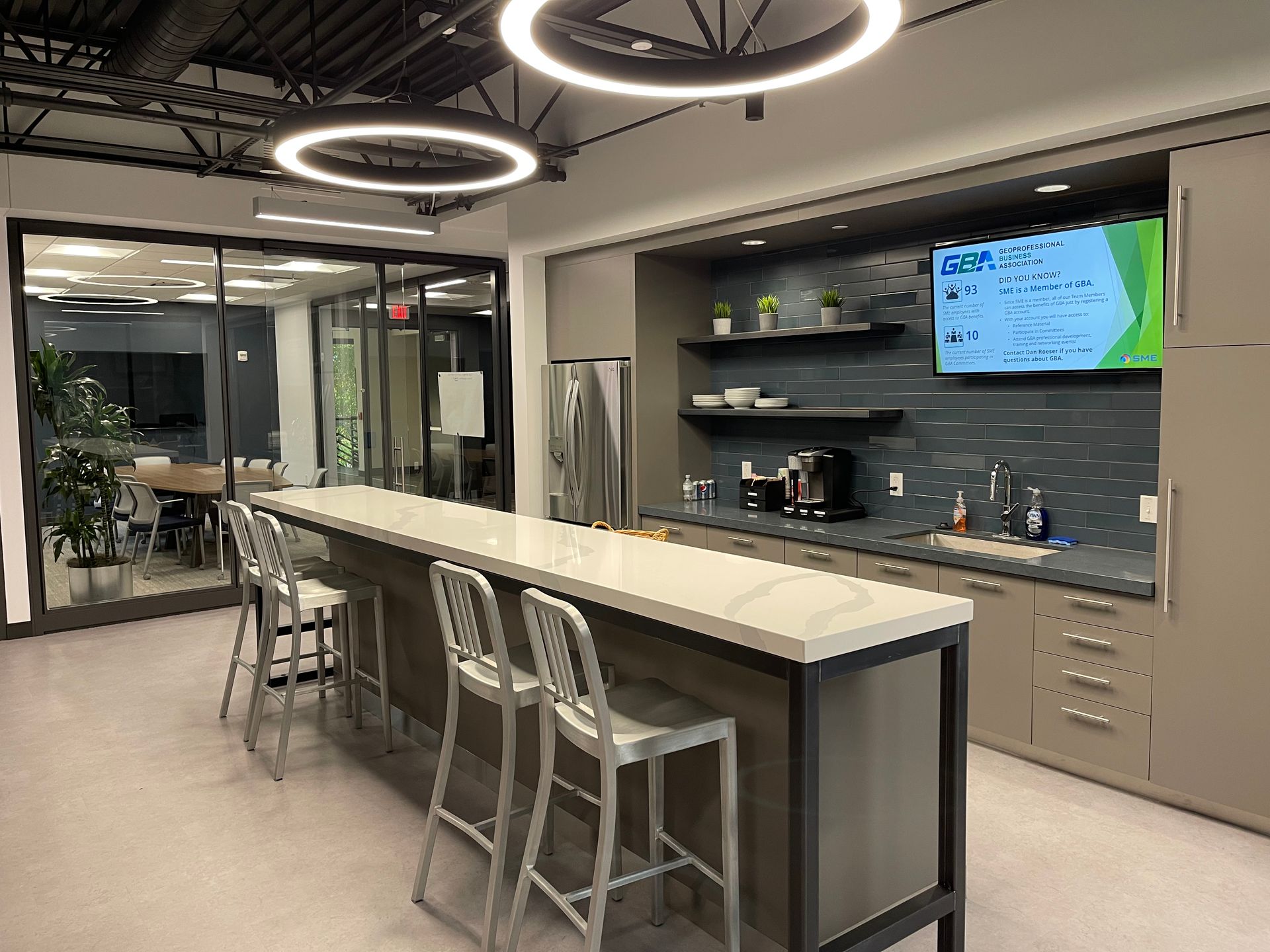 Modern office breakroom with island seating, kitchenette, and glass-walled conference room.