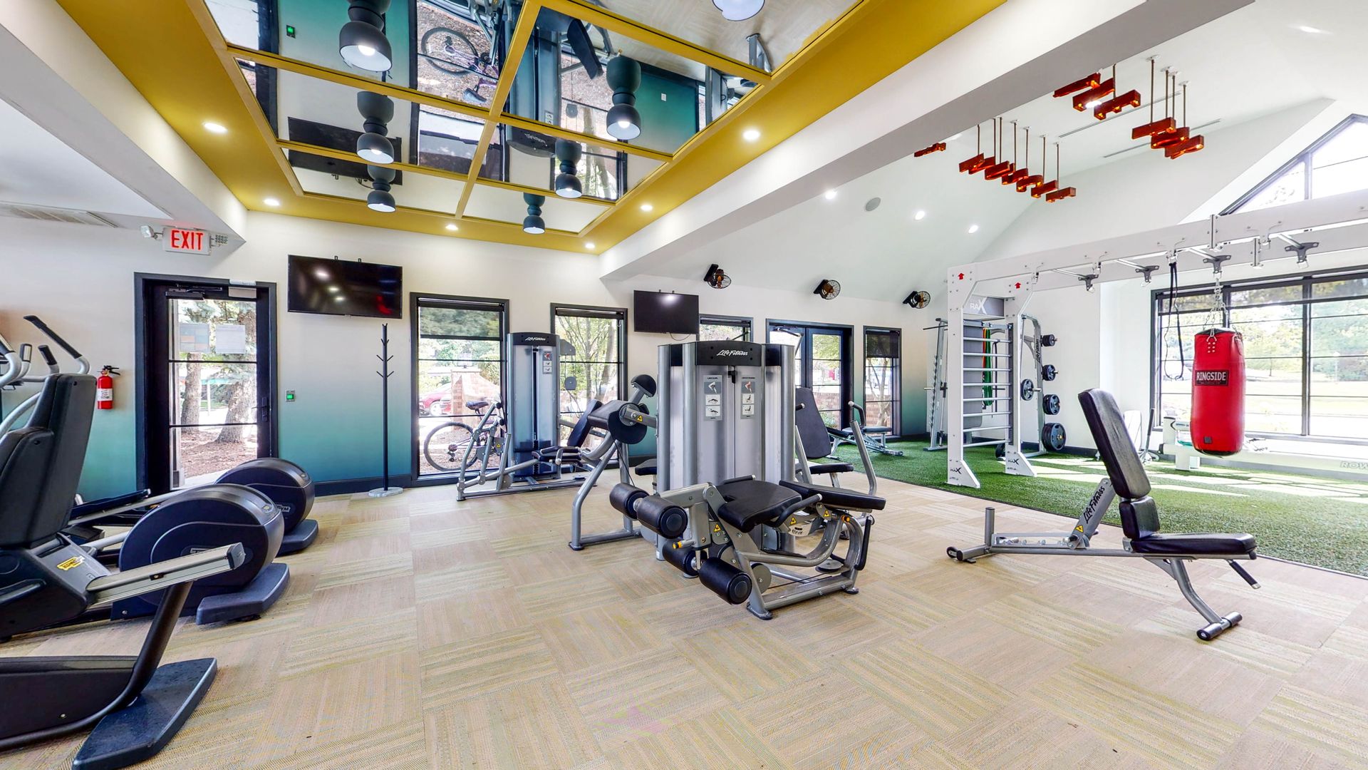 Gym with various exercise equipment, including machines, weights, and a punching bag. Green turf and mirrors.