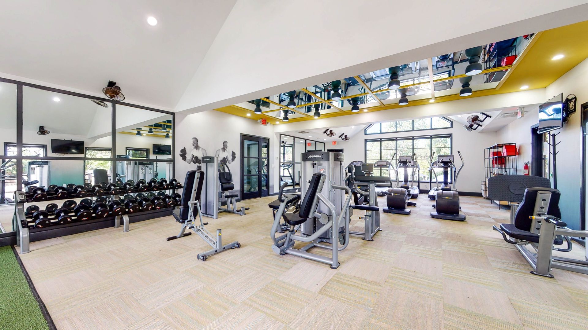 Gym interior with weights, machines, mirrors, and windows.