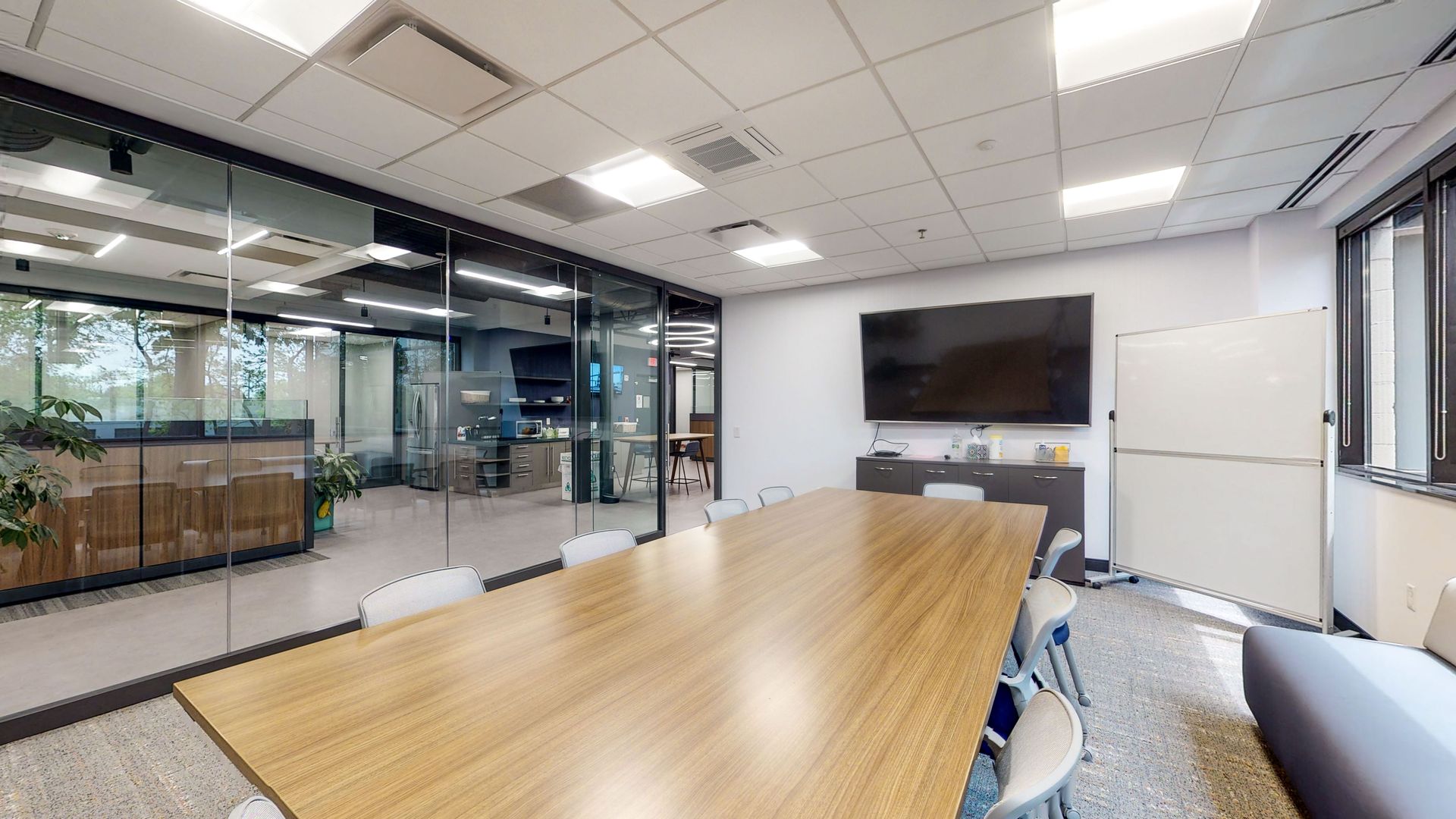 Conference room with large wooden table, chairs, and a TV on the wall. A glass wall overlooks an office area.