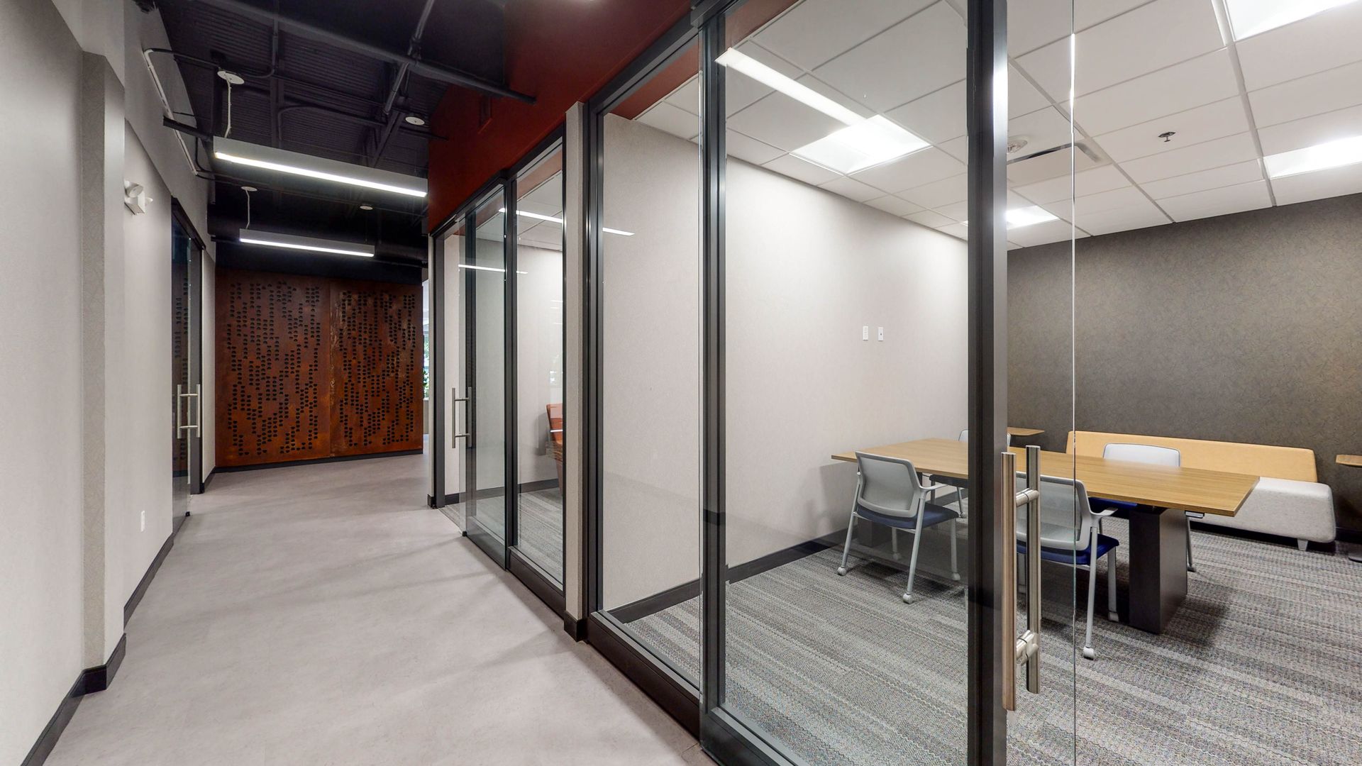 Corridor with glass-walled meeting rooms. One has a table and chairs, the other, a wood-paneled wall.
