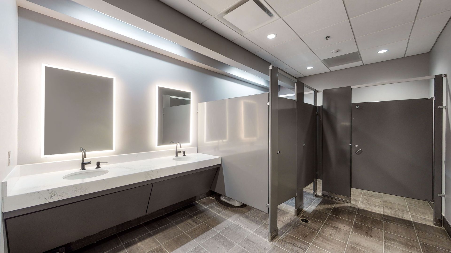 Modern, gray public restroom with two sinks, illuminated mirrors, and three stalls.