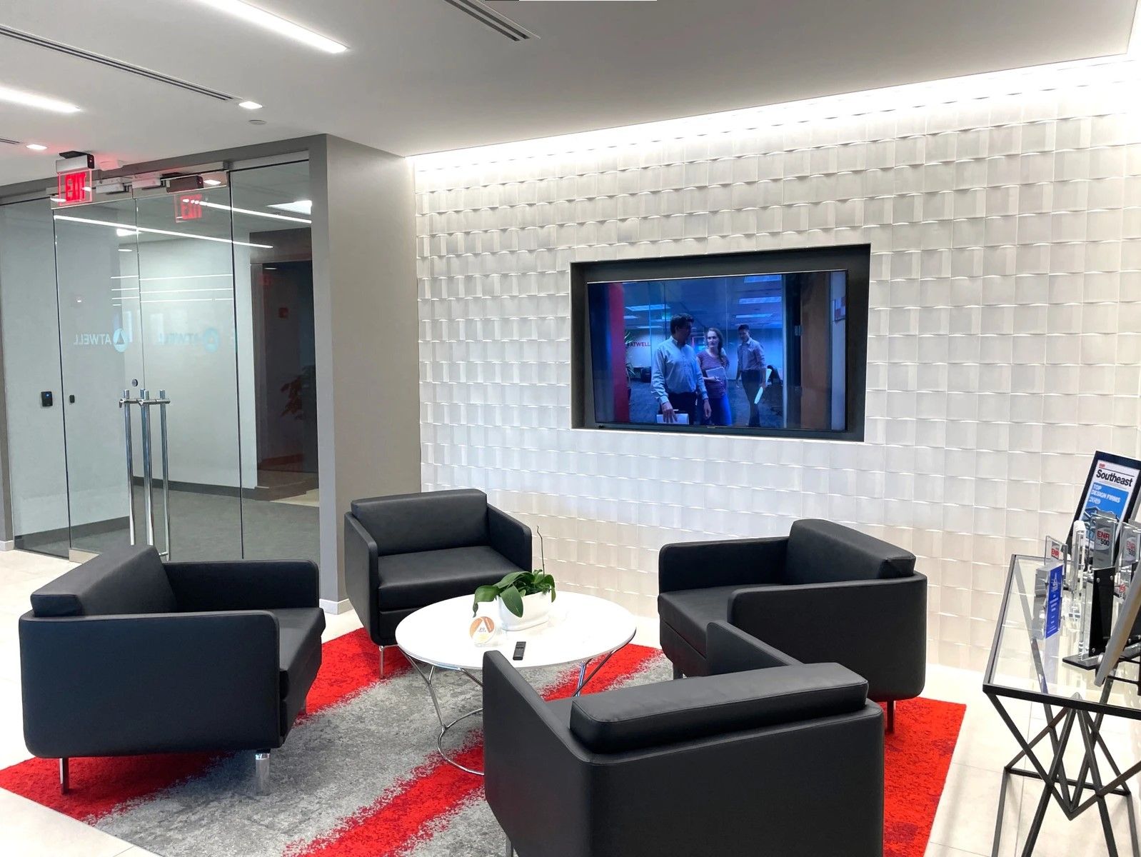 Modern lounge with black chairs, red rug, white table, and wall-mounted TV in a sleek office lobby