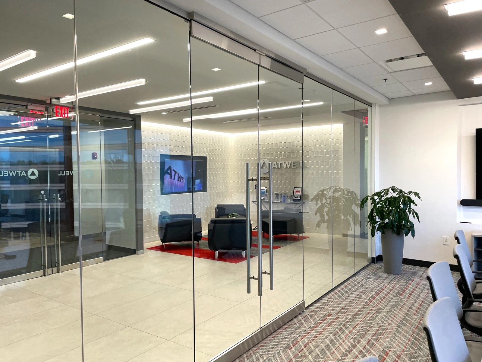 Modern office lounge with glass walls, red rug, black chairs, wall TV, and a potted plant
