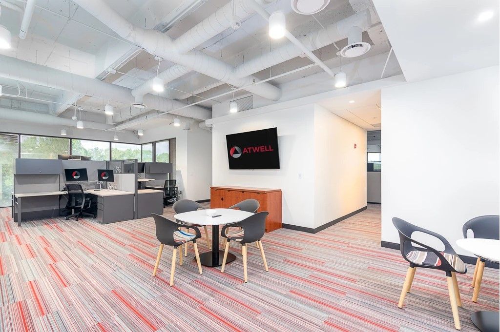 Modern office lounge with desks, chairs, round table, and wall-mounted TV in a bright open workspace