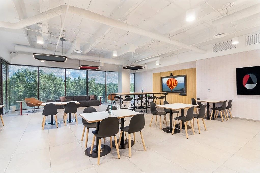 Modern office lounge with tables, chairs, large windows, and wall-mounted screens