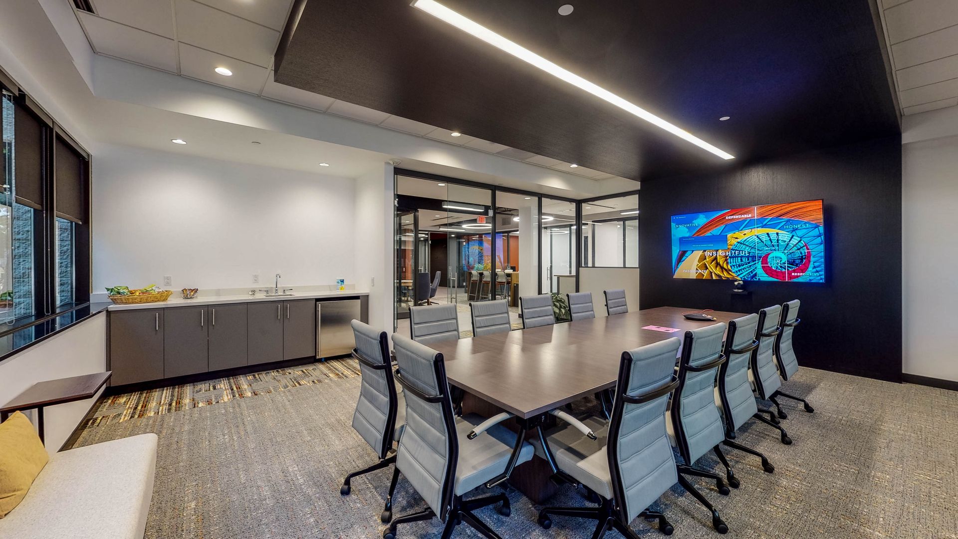 Modern conference room with long table, gray chairs, a large screen, and windows.