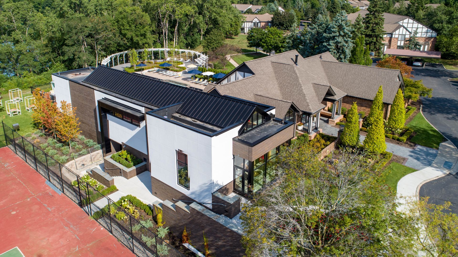 Aerial view of two connected houses, one white with a dark roof, the other brown with a brown roof, surrounded by greenery.