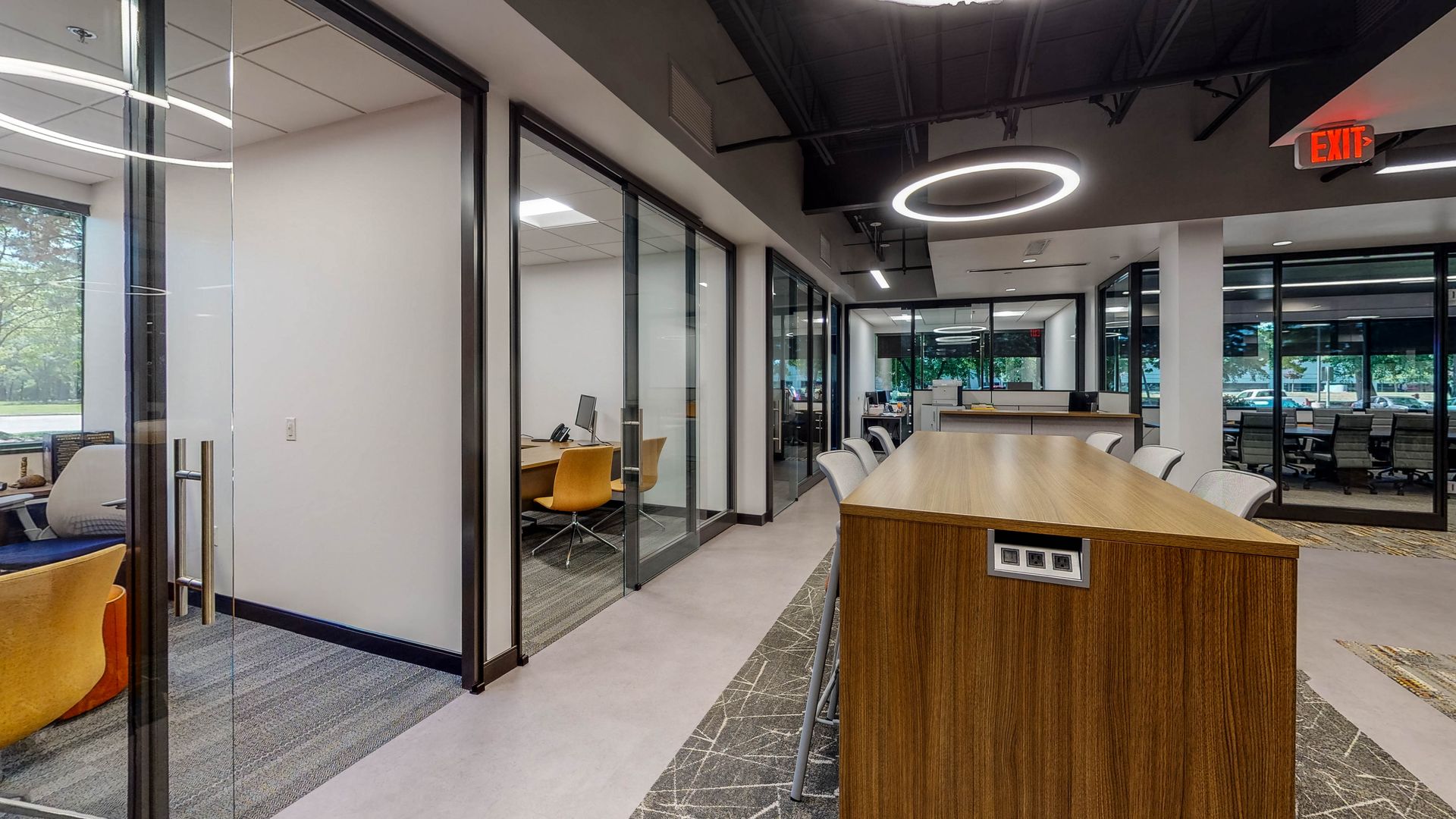 Modern office hallway with glass-walled offices, long wooden table, and circular lights.