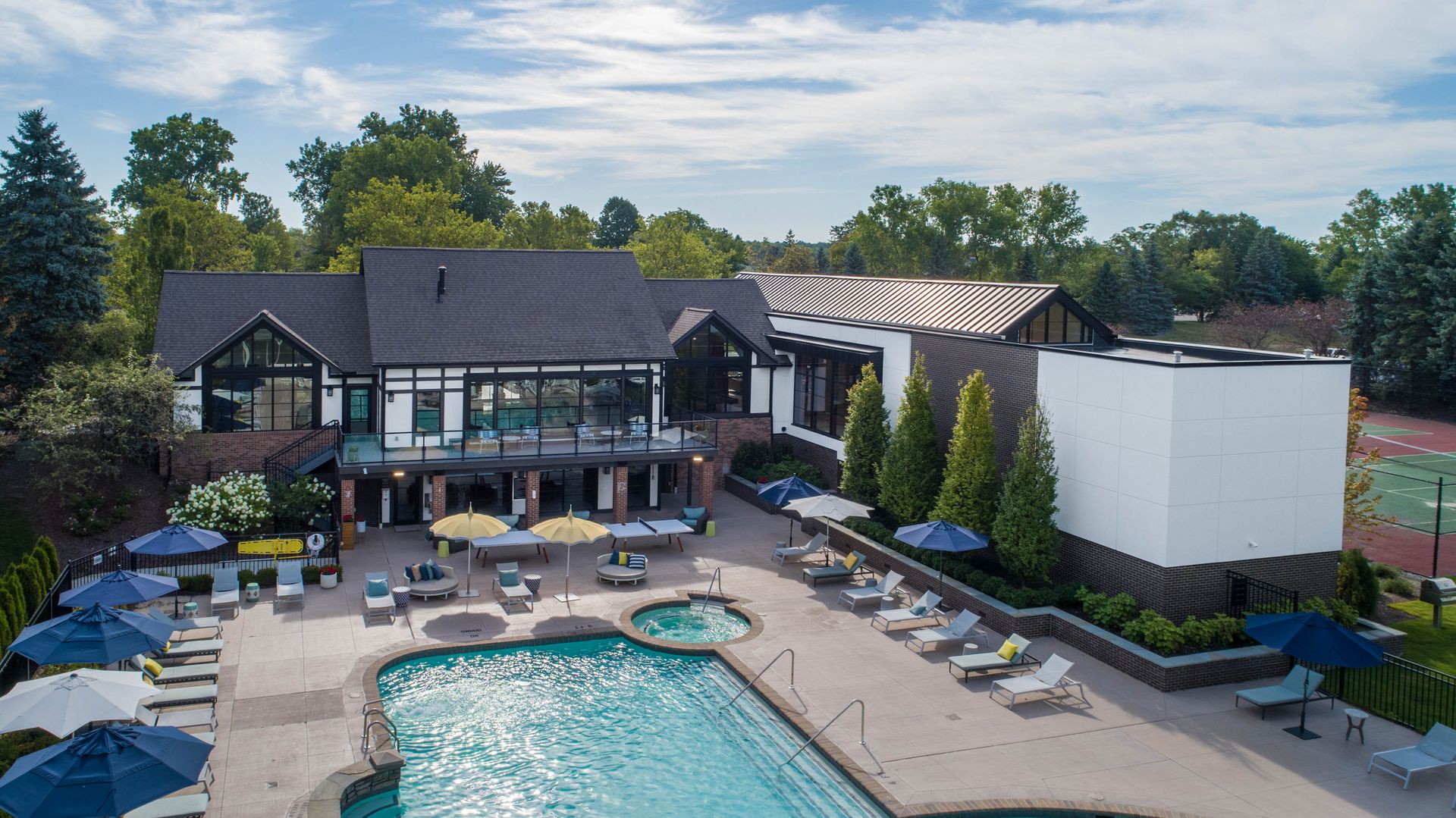 Aerial view of a club with a pool, lounge chairs, and tennis court. The building is a mix of styles.