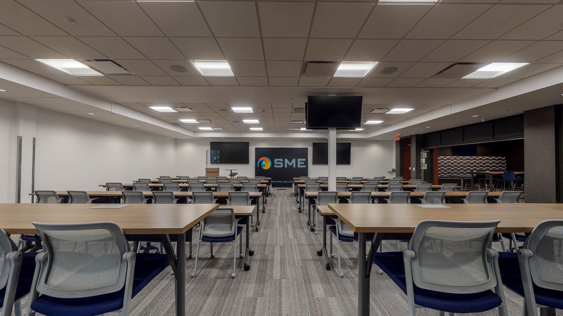 Classroom with rows of tables and chairs, screens, and a logo on the wall.