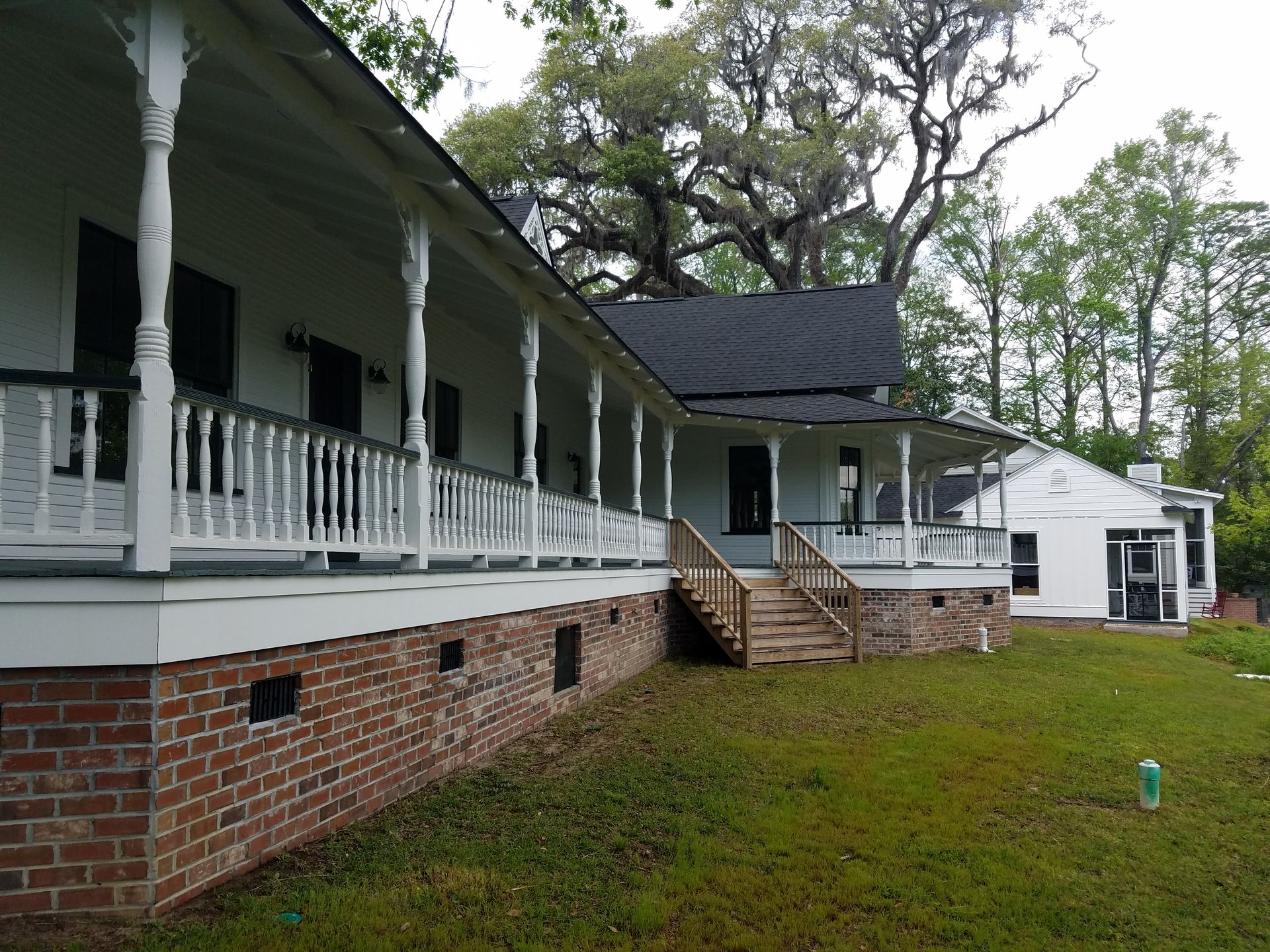 White house with wrap-around porch, brick foundation, and black shutters. Green lawn, trees in the background.