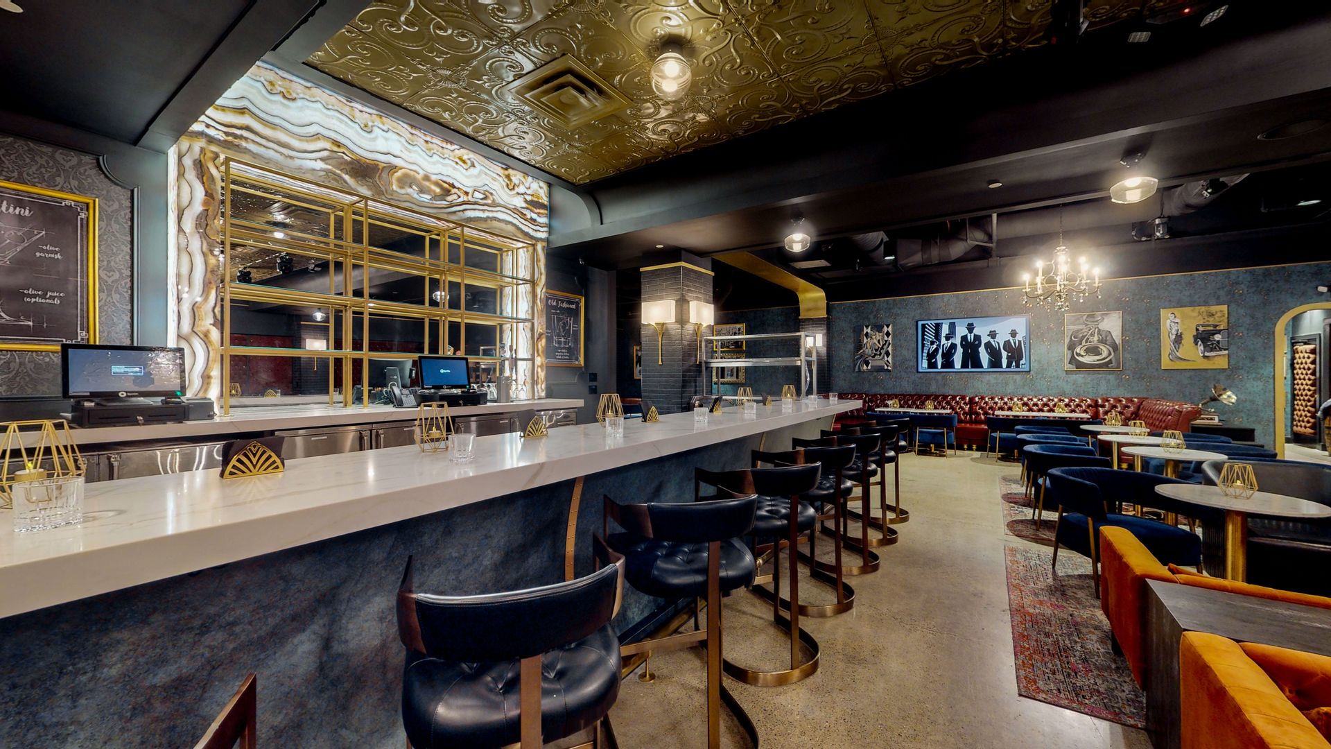 Bar interior with gold and marble accents. Black chairs line a long bar. Tables and seating area in the back.