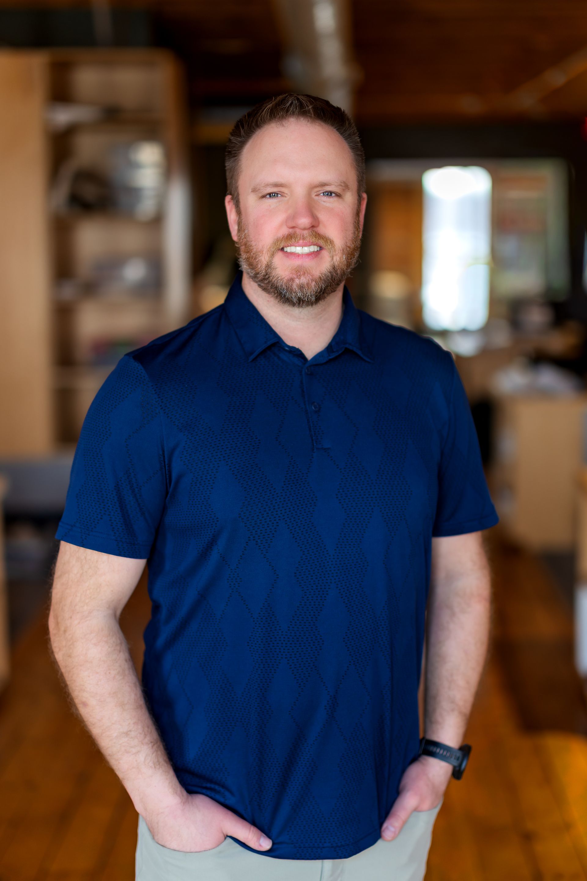 Man with a beard smiles at the camera, wearing a blue patterned shirt, neutral background.