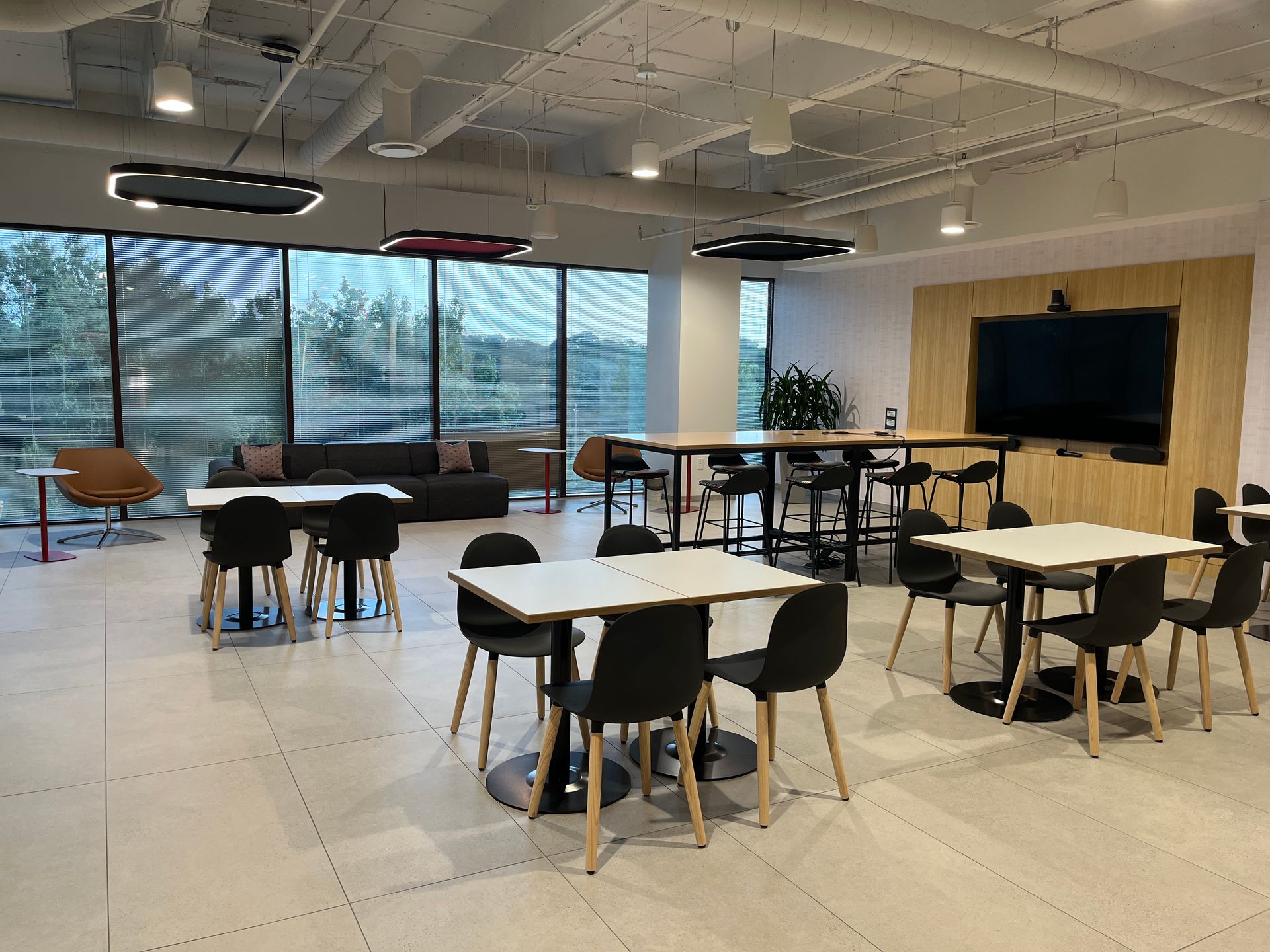 Modern lounge with tables, chairs, large windows, and a wall-mounted TV in a bright open room