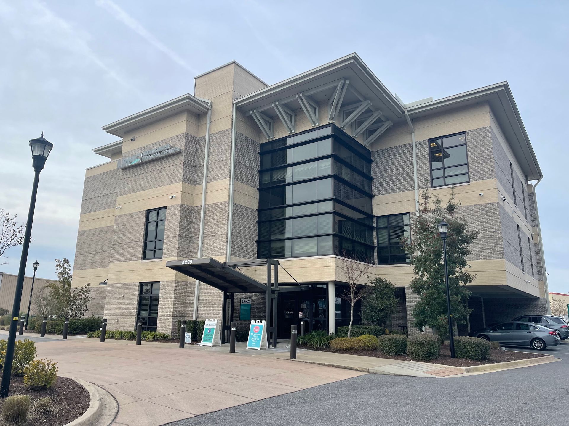 Beige brick building with a glass entrance and parking in front. Cloudy sky.