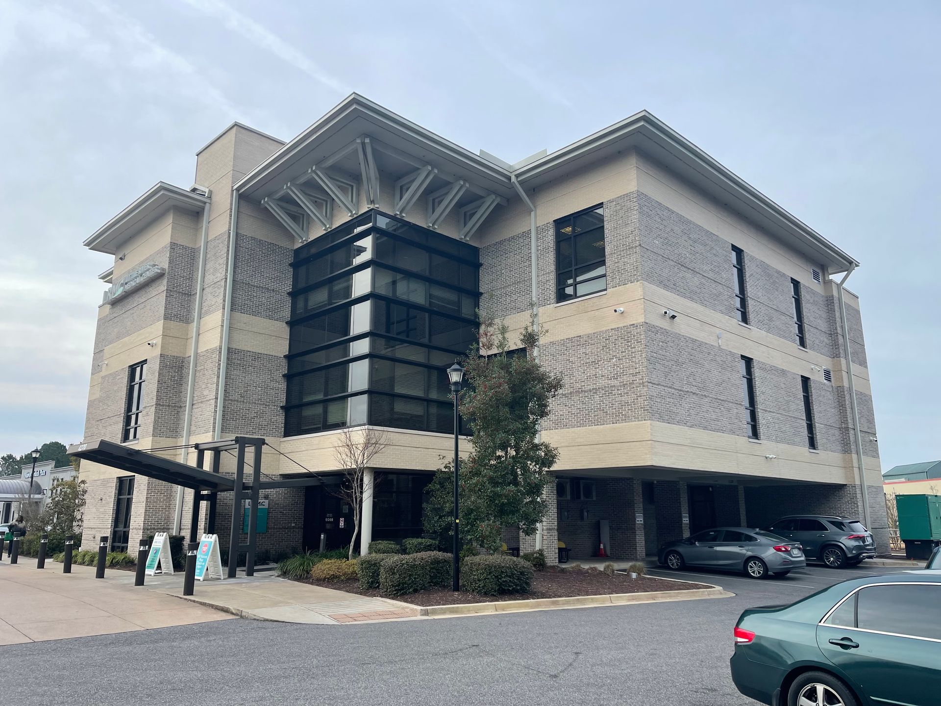 Modern three-story building with textured beige walls, a black glass section, and a covered entrance; cars parked nearby.