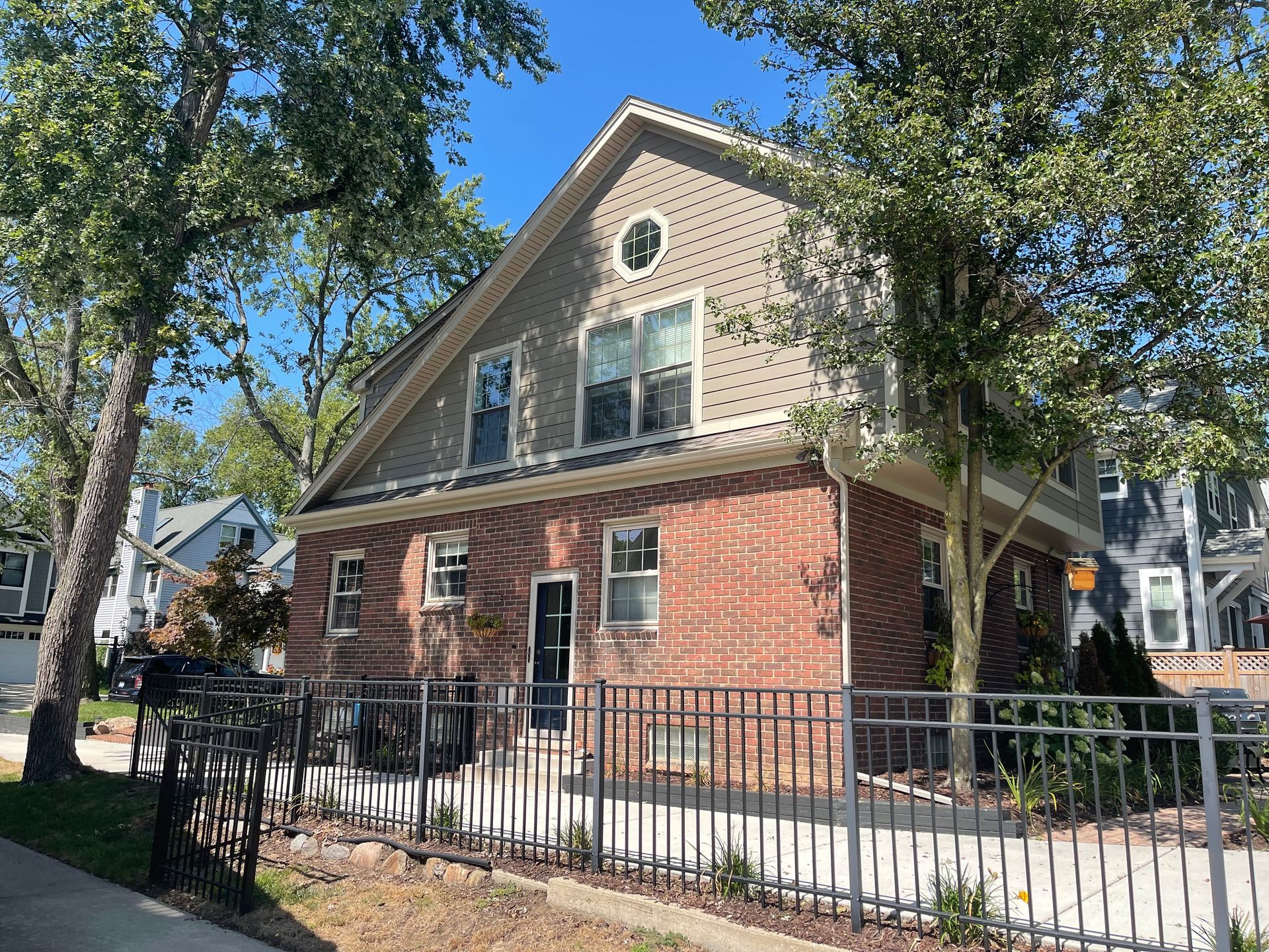 Two-story brick house with a gray gable, dark fence, and trees on a sunny day.