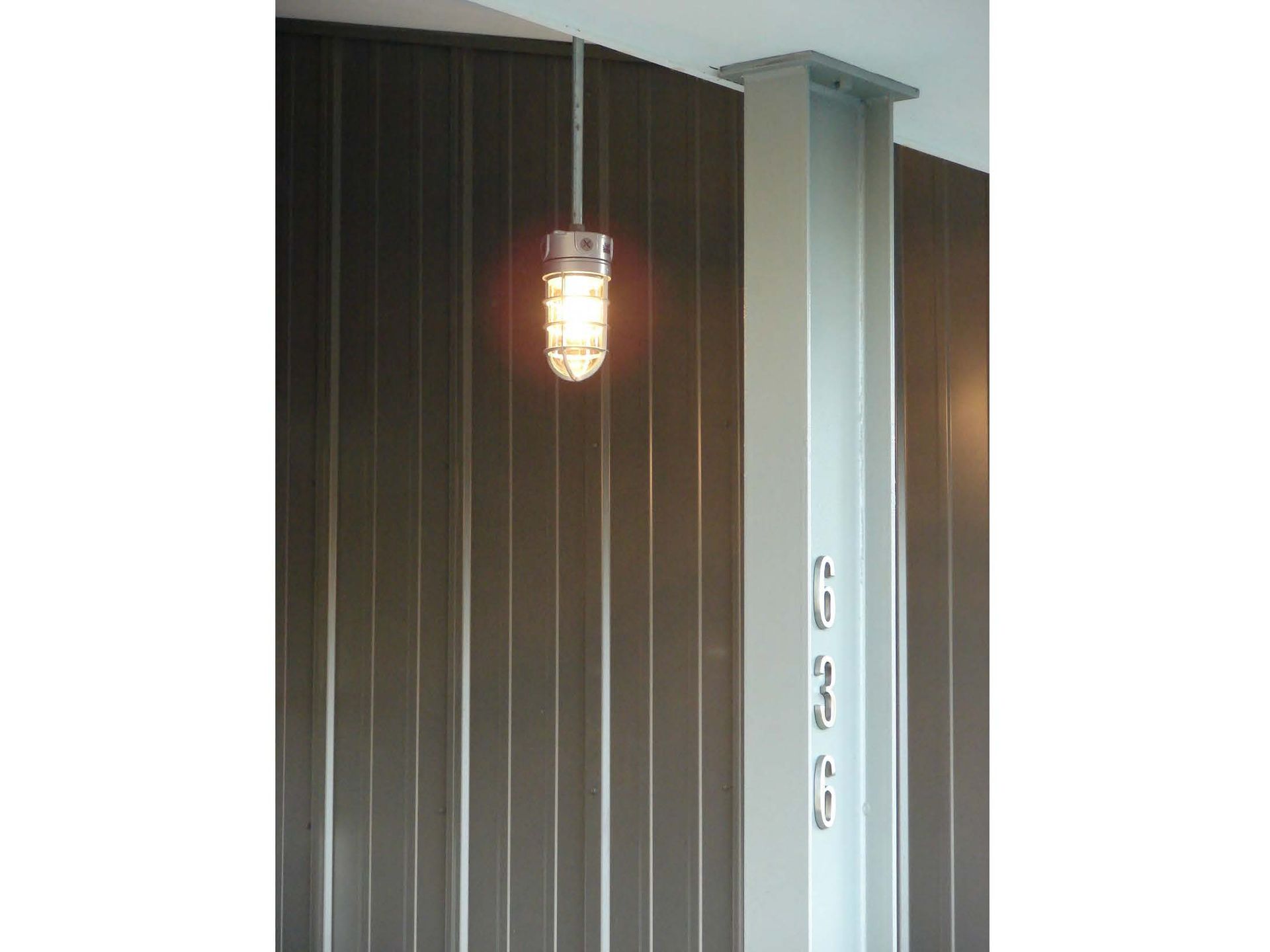 Industrial-style hanging light fixture with a caged glass bulb illuminates a brown corrugated wall near a white structural beam.