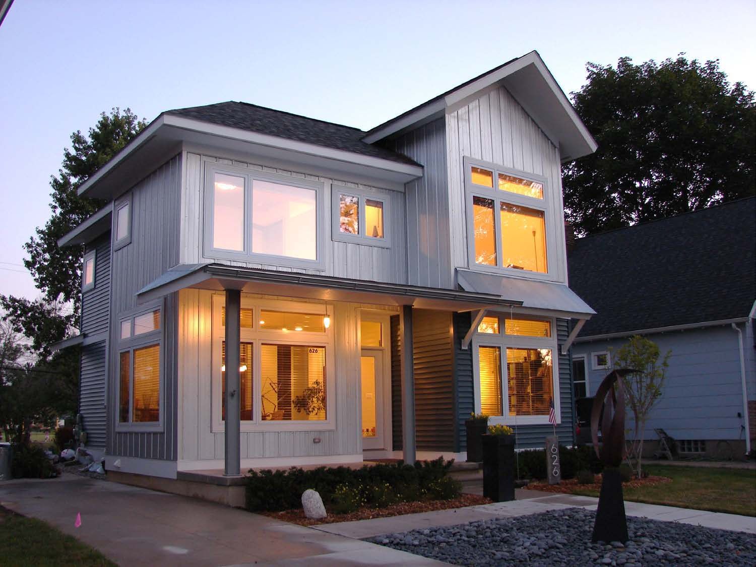 Modern two-story house with light blue siding, large windows, and a porch. Lit interior.