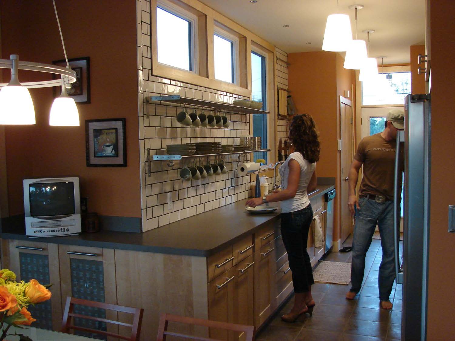 Kitchen with two people: one cooking, one near fridge. Orange walls, light wood cabinets.