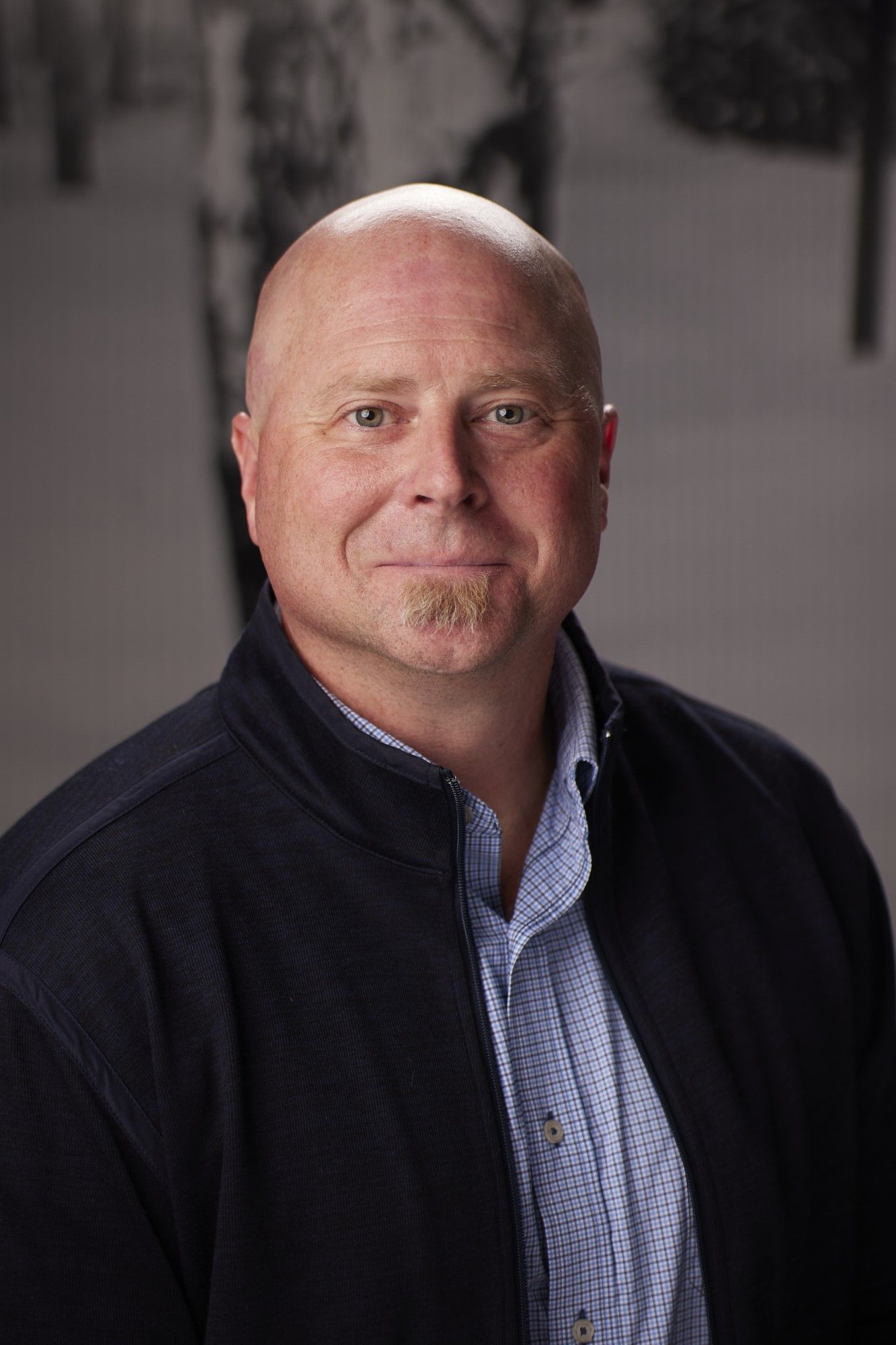 Bald man, goatee, wearing a blue jacket over a button-down shirt, smiling. Background is blurred.