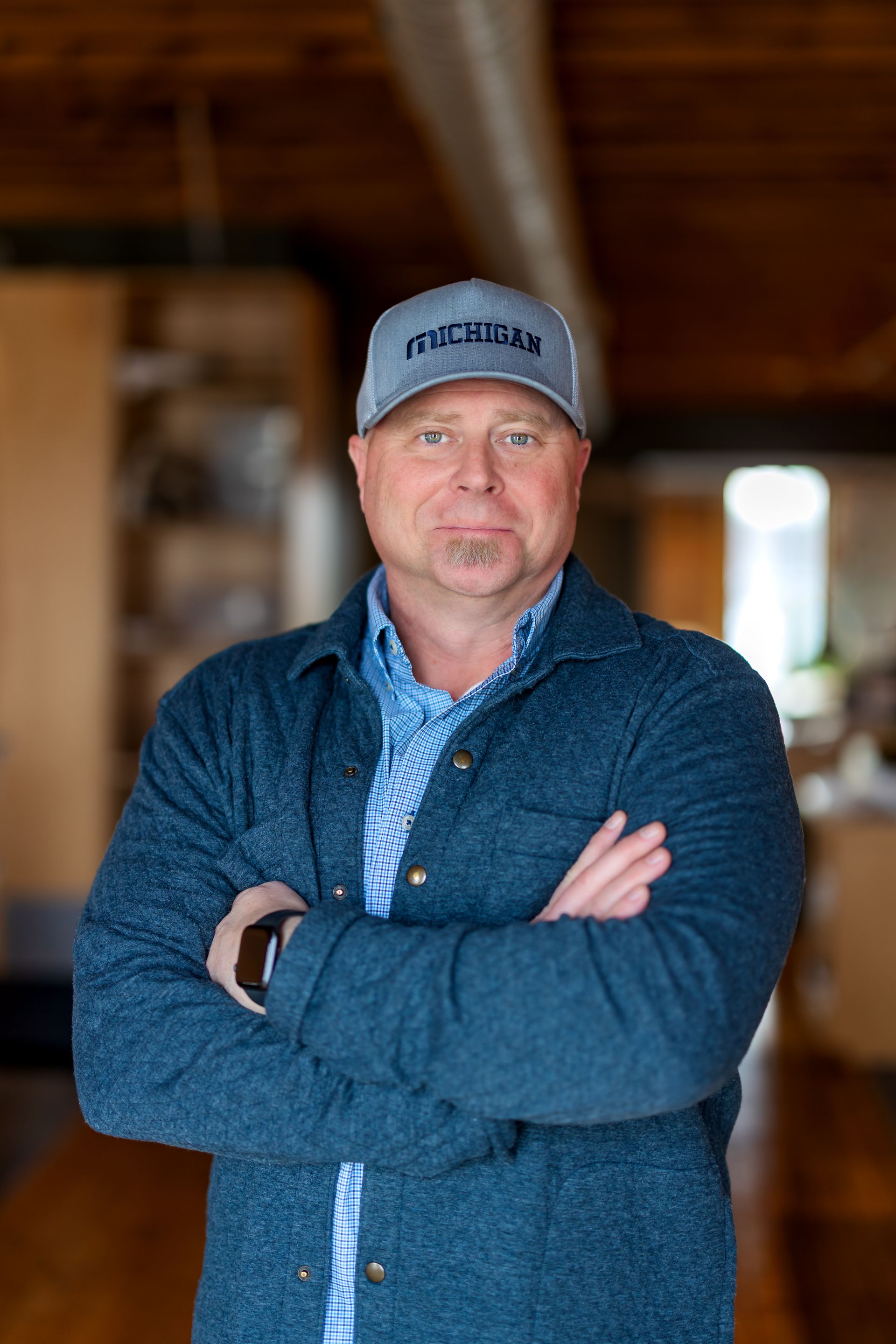 Bald man, goatee, wearing a blue jacket over a button-down shirt, smiling. Background is blurred.
