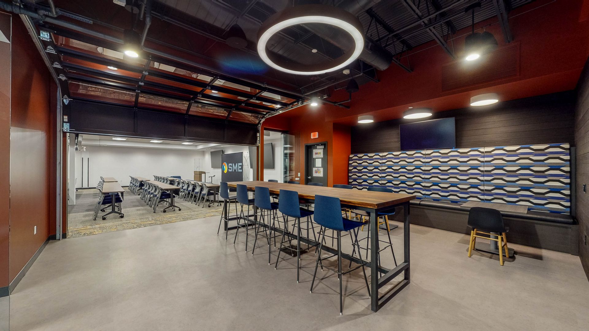 Modern office lounge with high tables, blue stools, and a circular ceiling light in an industrial space