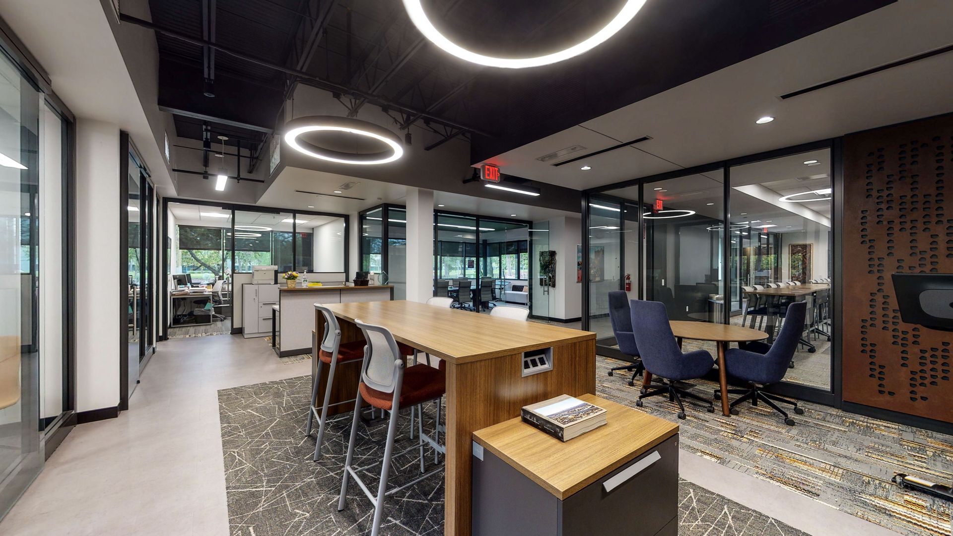 Modern office lounge with wooden table, stools, sofas, and glass-walled meeting rooms