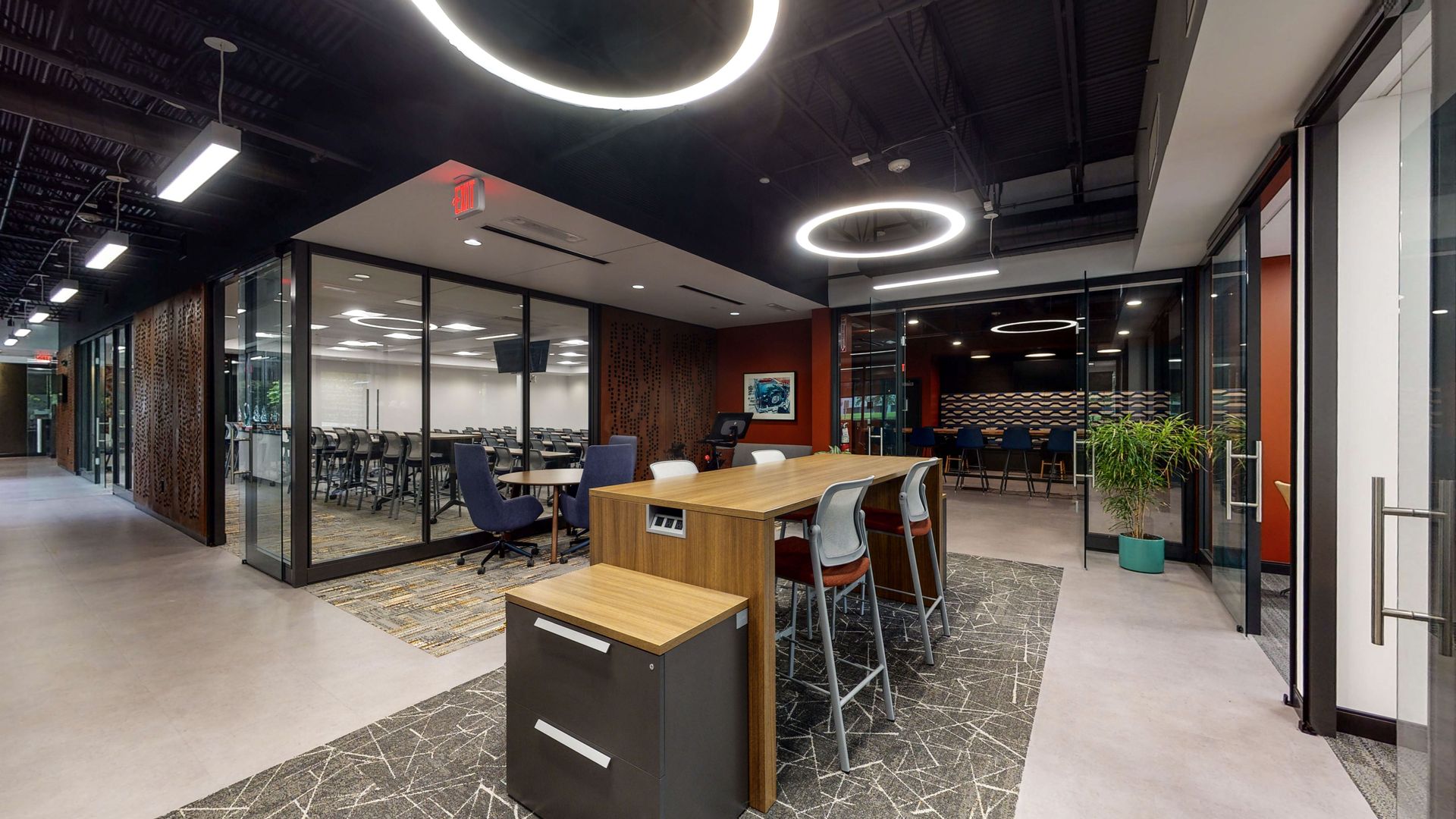 Modern office lounge with bar stools, tables, glass-walled meeting rooms, and circular ceiling lights