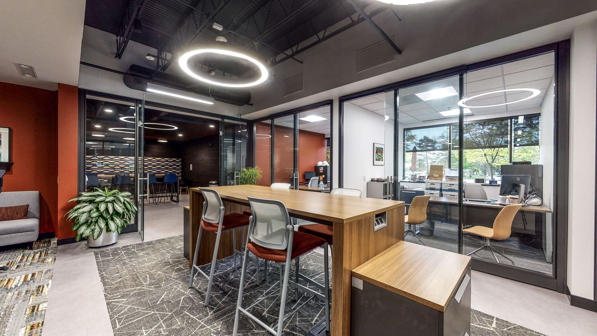 Modern office lounge with wooden tables, stools, glass-walled meeting rooms, and a ring light fixture