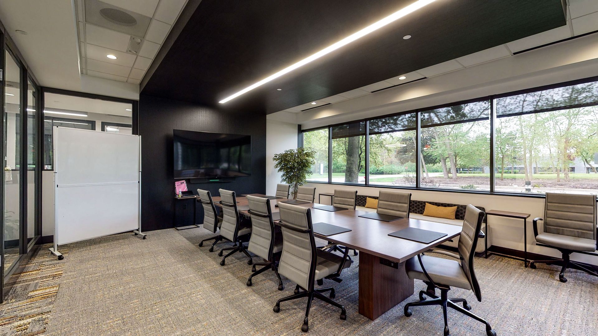 Modern conference room with long table, rolling chairs, wall-mounted TV, large windows, and whiteboard