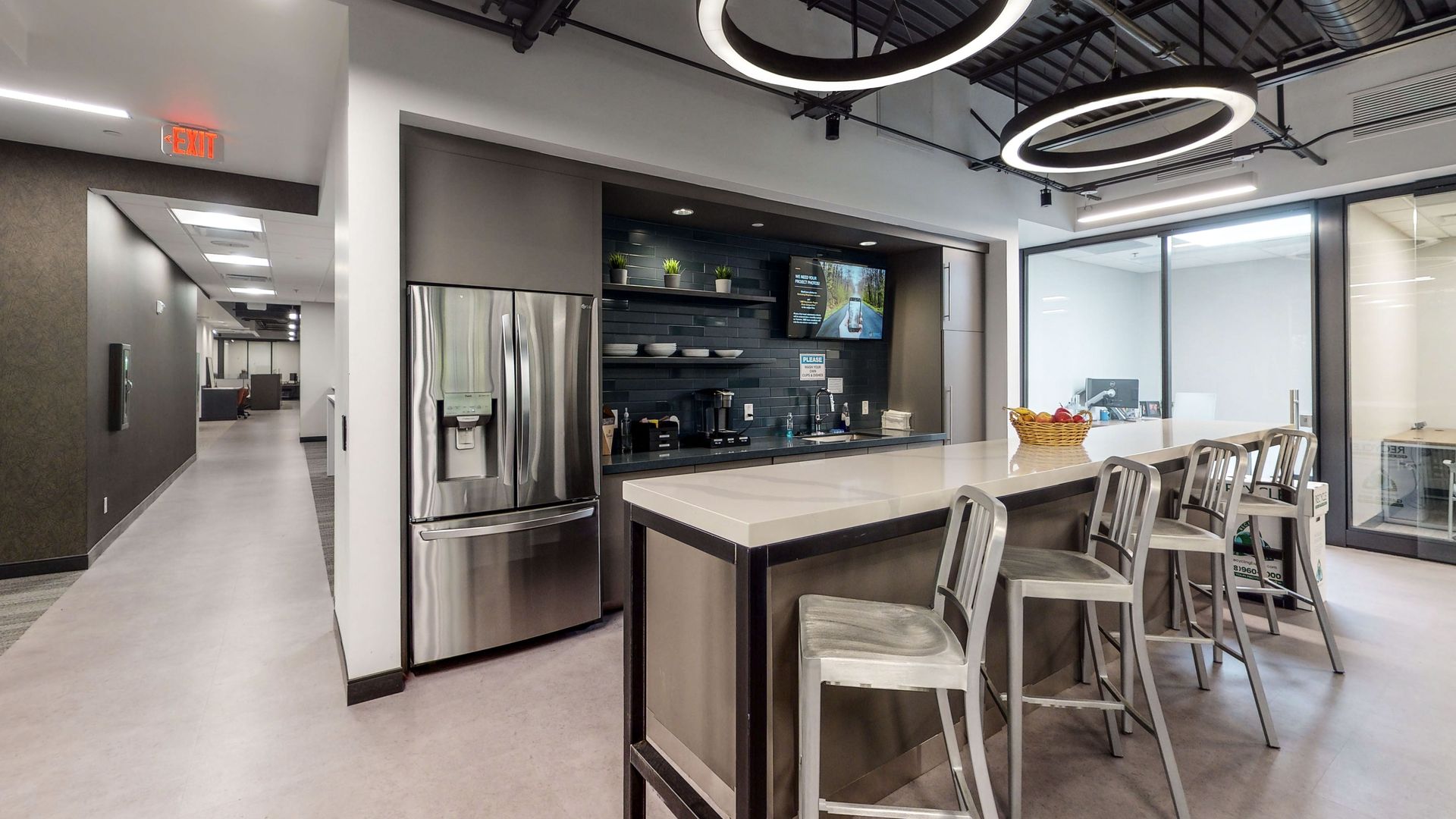 Modern office break room with stainless steel fridge, island table, stools, and ring ceiling lights