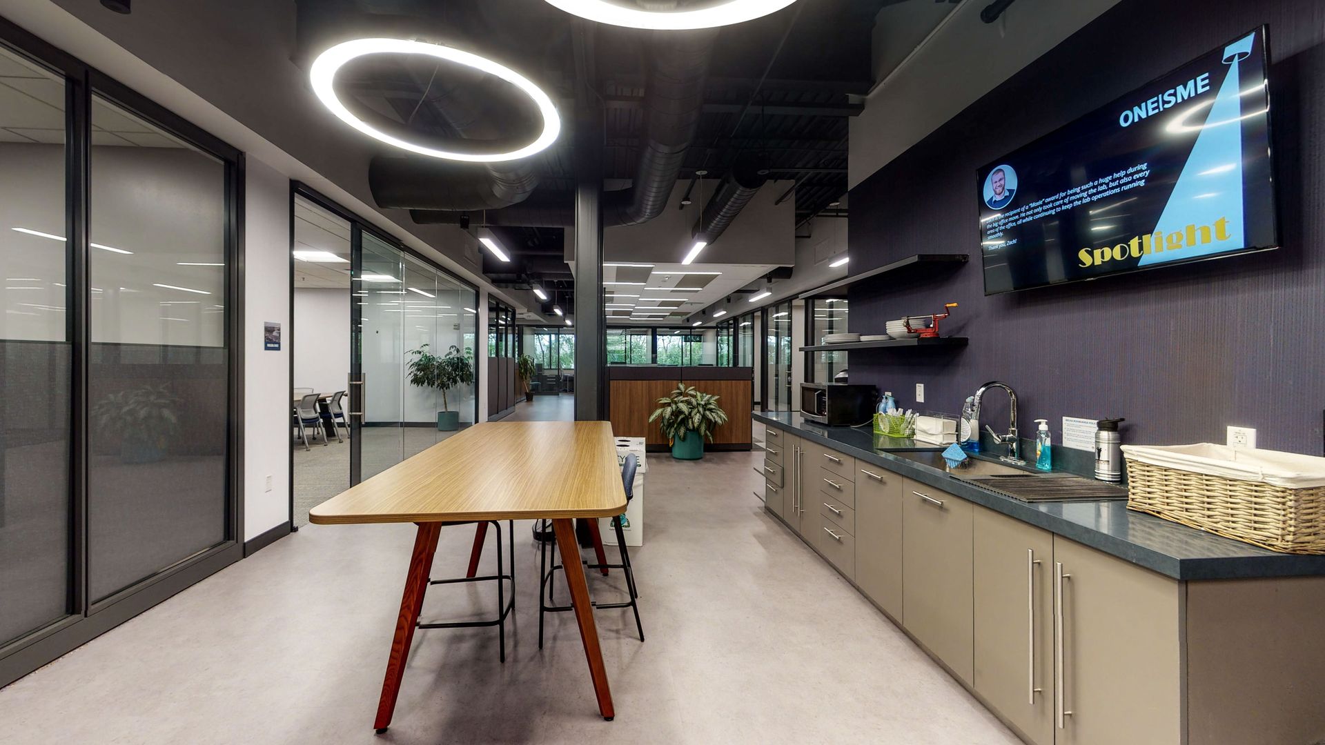 Modern office kitchenette with a long wooden table, cabinets, chairs, and a wall-mounted display on the right.