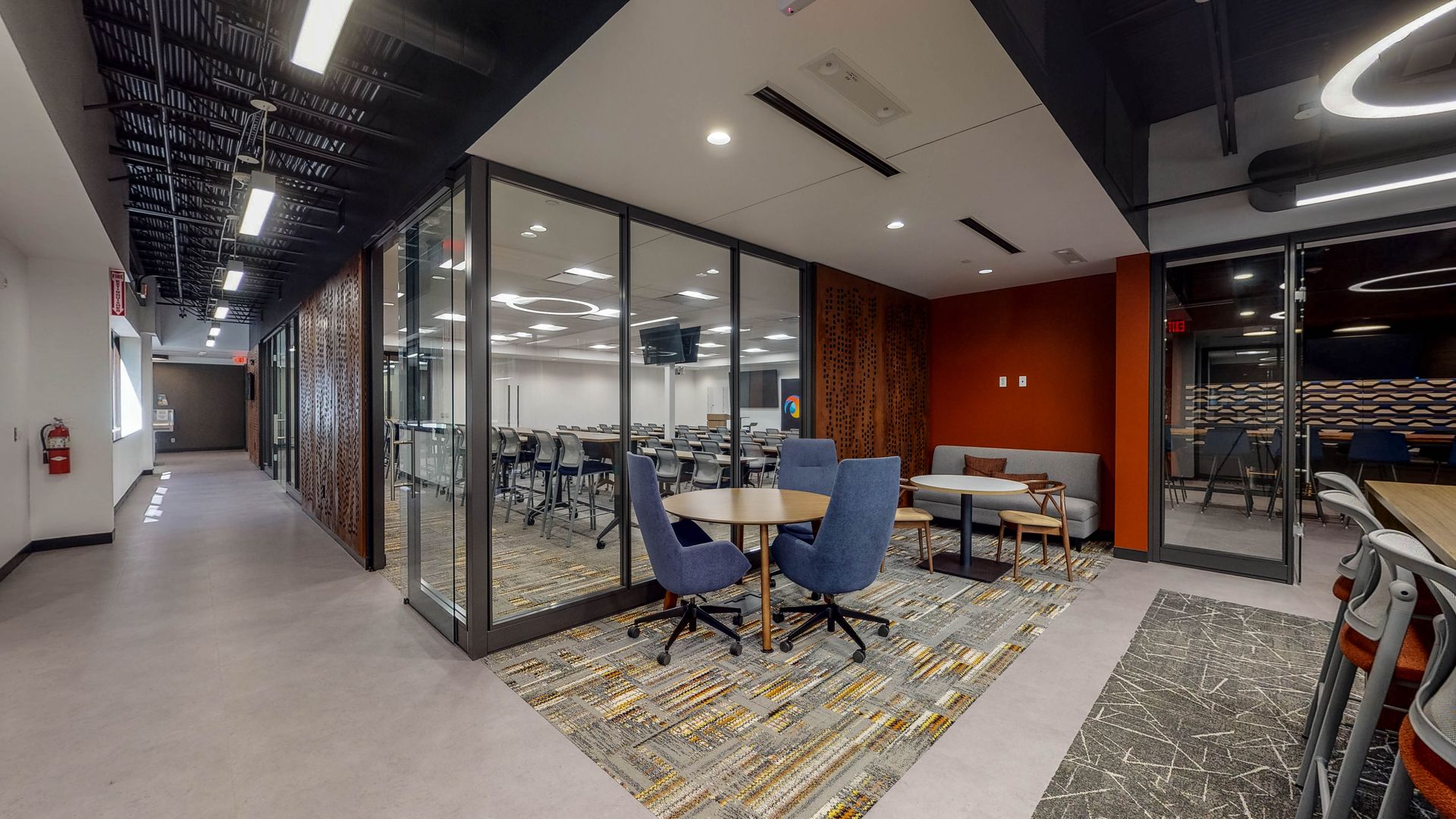 Modern office lounge with glass-walled meeting room, round tables, chairs, and patterned rugs in a corridor