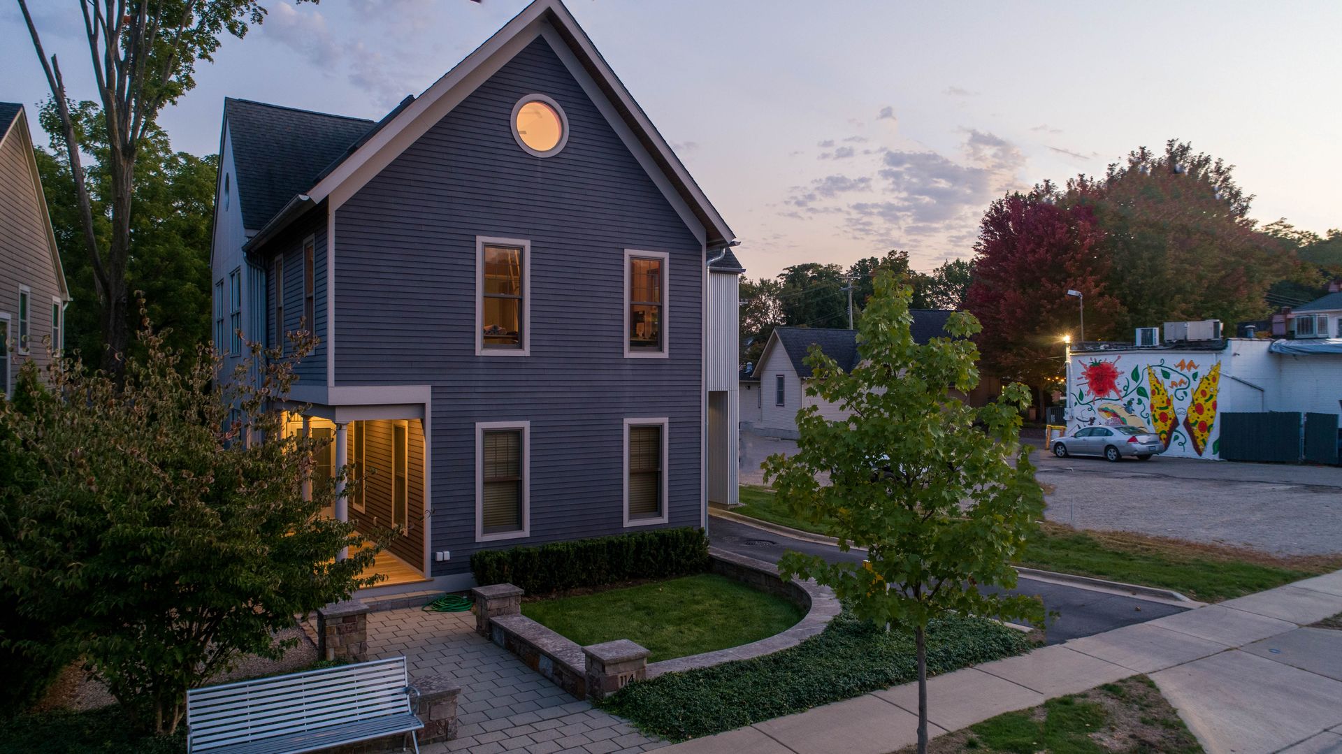 A gray two-story house with a circular window. It's in a neighborhood setting at dusk, with greenery and a mural.