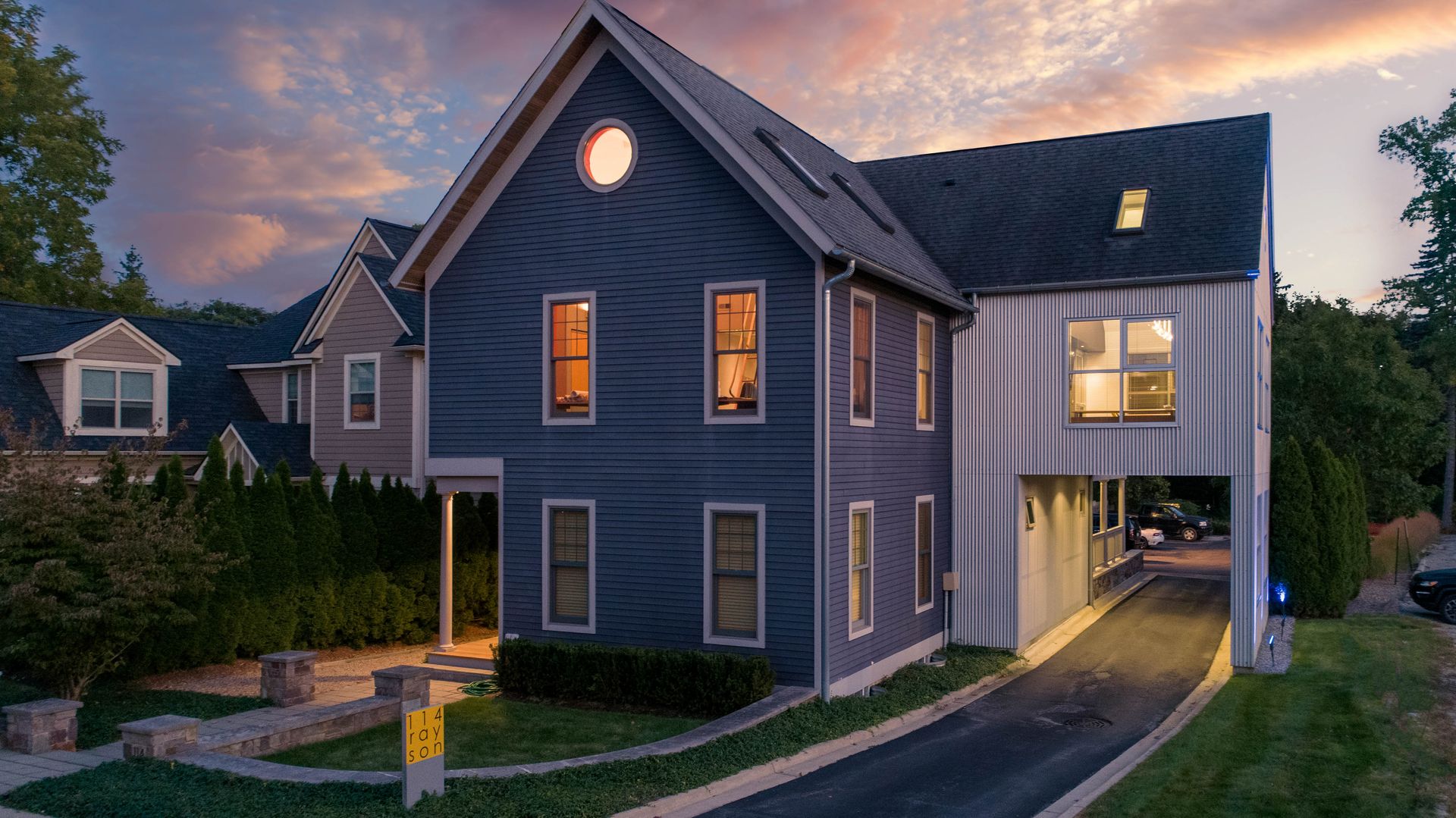 Two-story blue house with a driveway, a connecting structure and round window at dusk.