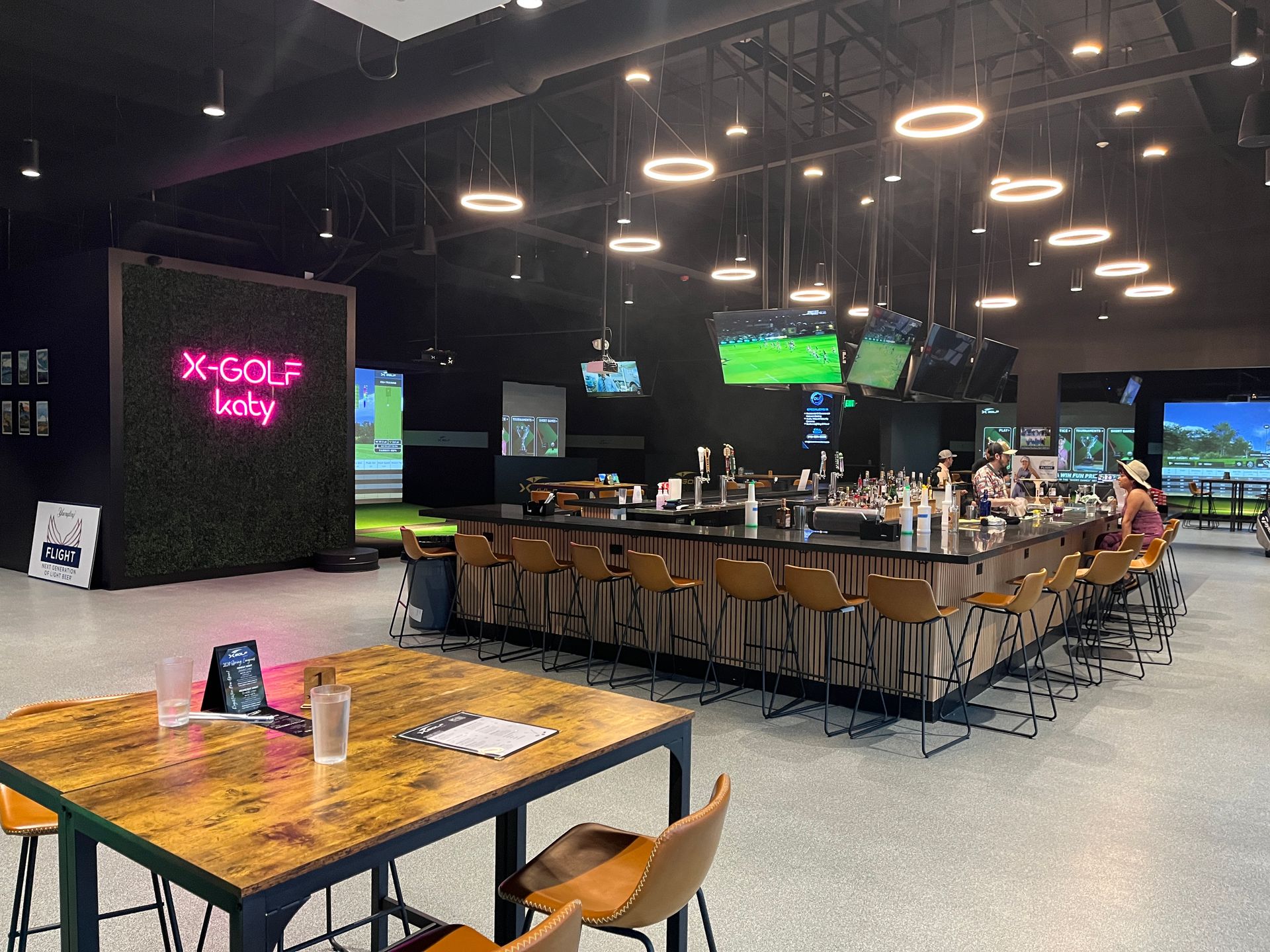 Interior of X-Golf bar with a central bar, tables, golf simulators, TVs, and circular lighting.