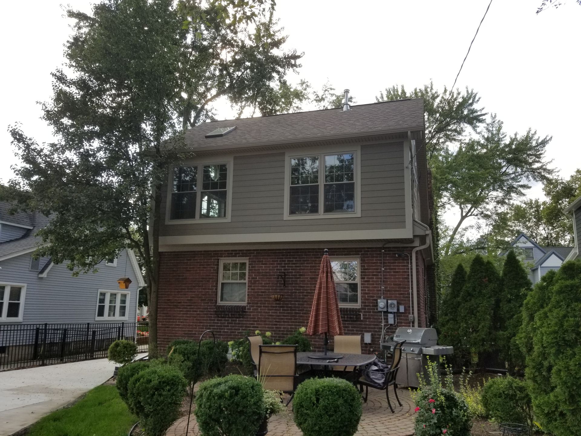 Back of two-story house with brick bottom, gray siding top, windows, patio furniture, and surrounding greenery.